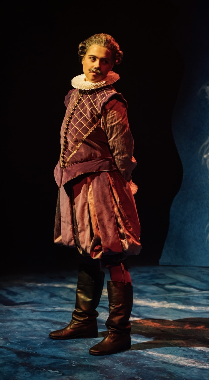 Actor in Elizabethan costume with burgundy doublet, white ruff collar, and leather boots on stage with blue lighting.
