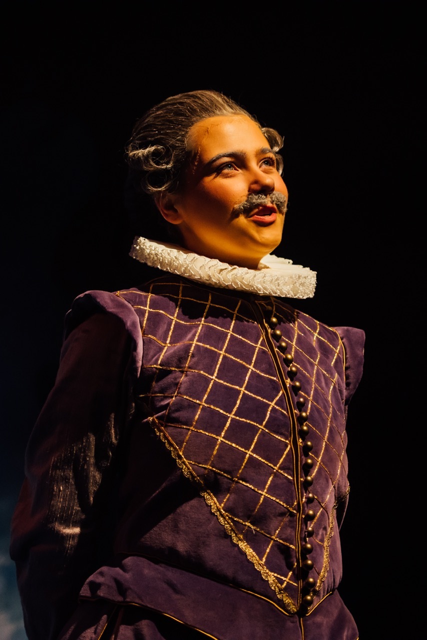 Actor in purple Elizabethan doublet with gold diamond pattern, white ruff collar, and period moustache on dark stage.
