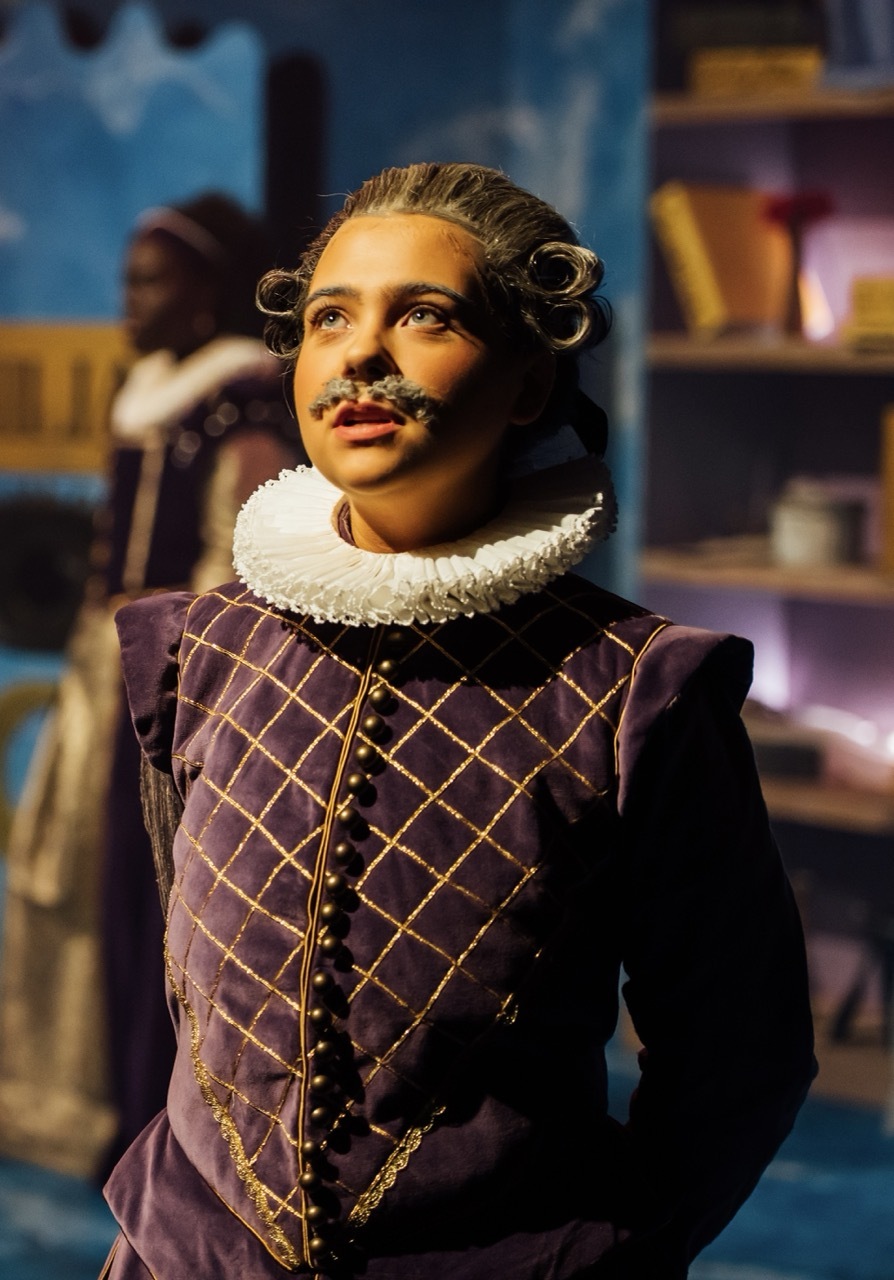 Actor in Elizabethan costume with purple doublet, gold diamond pattern, white ruff collar, and curled moustache on theatre stage.