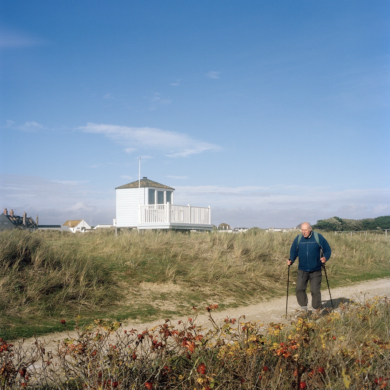 An older man walking with trekking poles along a sandy path, with dunes and a white beach hut under a clear blue sky.