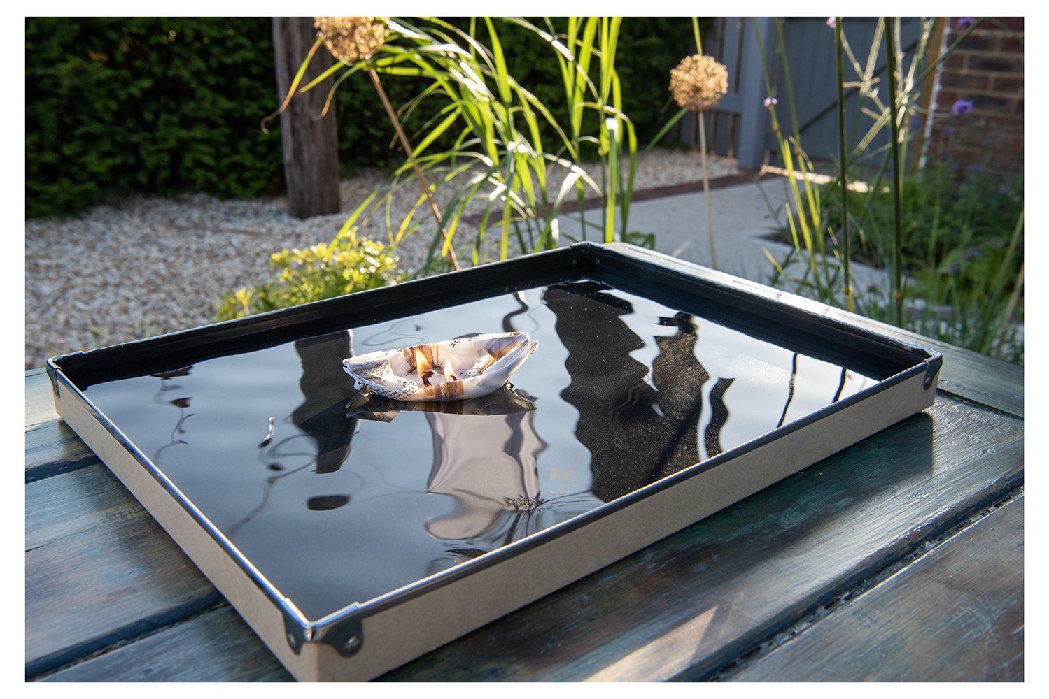 Paper boat floating on a black shallow tray of water so it acts like a mirror, reflecting my garden, clothes hanging on the washing line and plants/flowers growing.