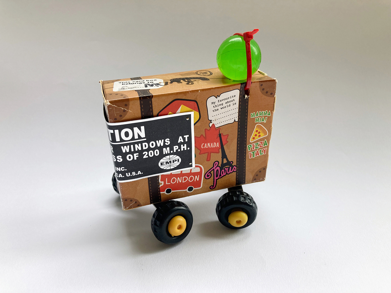 Cardboard box printed with suitcase design. 4 plastic wheels attached to box, sticker windscreen and bouncy ball luggage.