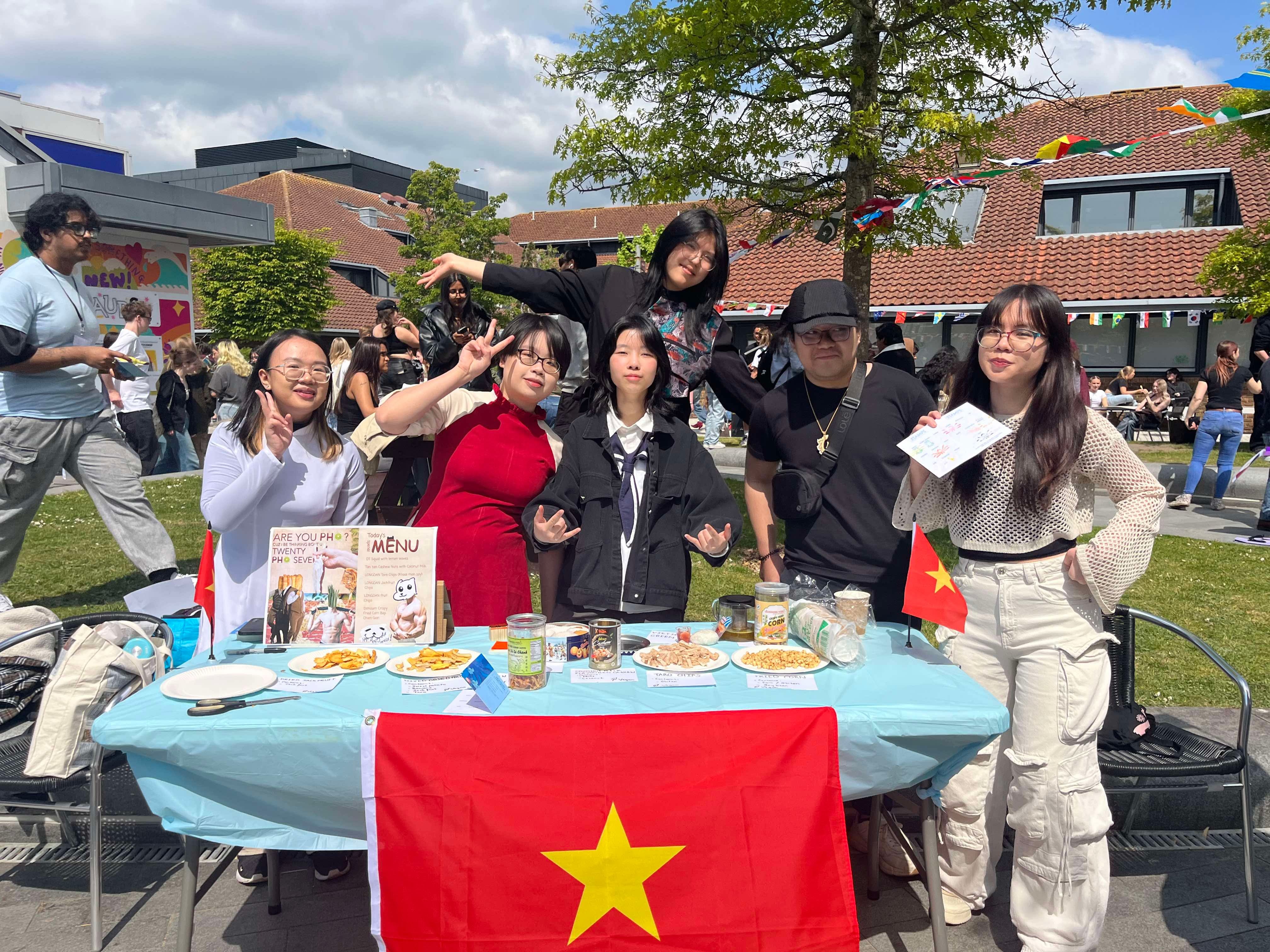 Group of six people posing at outdoor event table with Vietnamese flag, food displays, and brick buildings in background.