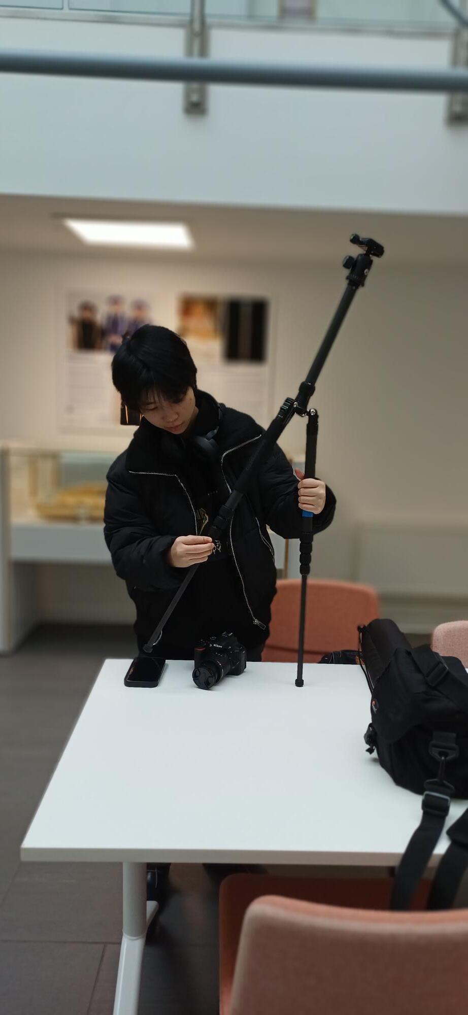 Person in black jacket kneeling on white table, holding camera equipment in modern indoor space with orange chairs and white walls.
