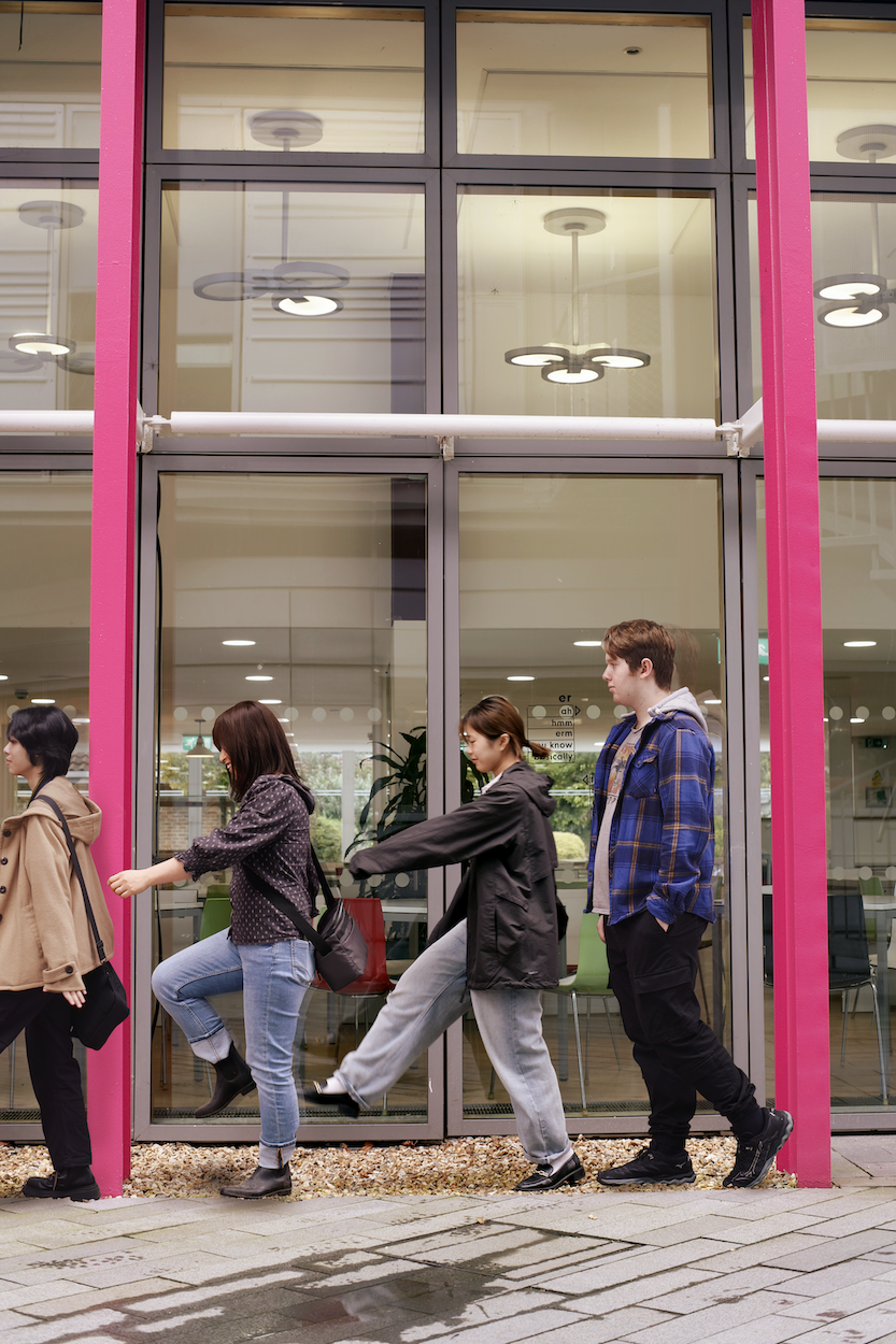 Four young people entering a modern building through glass doors with bright pink frames on a paved walkway.