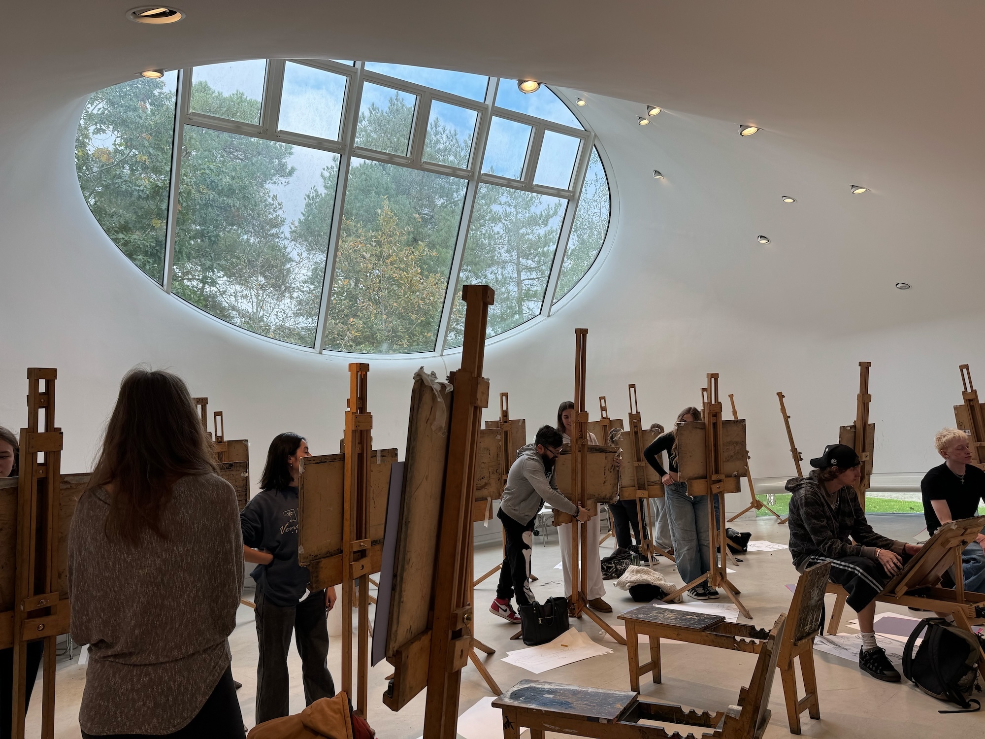Art studio with students at wooden easels beneath circular skylight showing trees outside. White walls, recessed lighting.