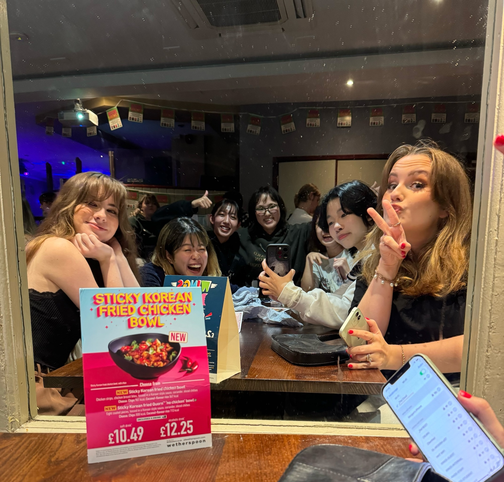 Group of people at restaurant table making peace signs, with Korean fried chicken bowl menu card displayed in foreground.