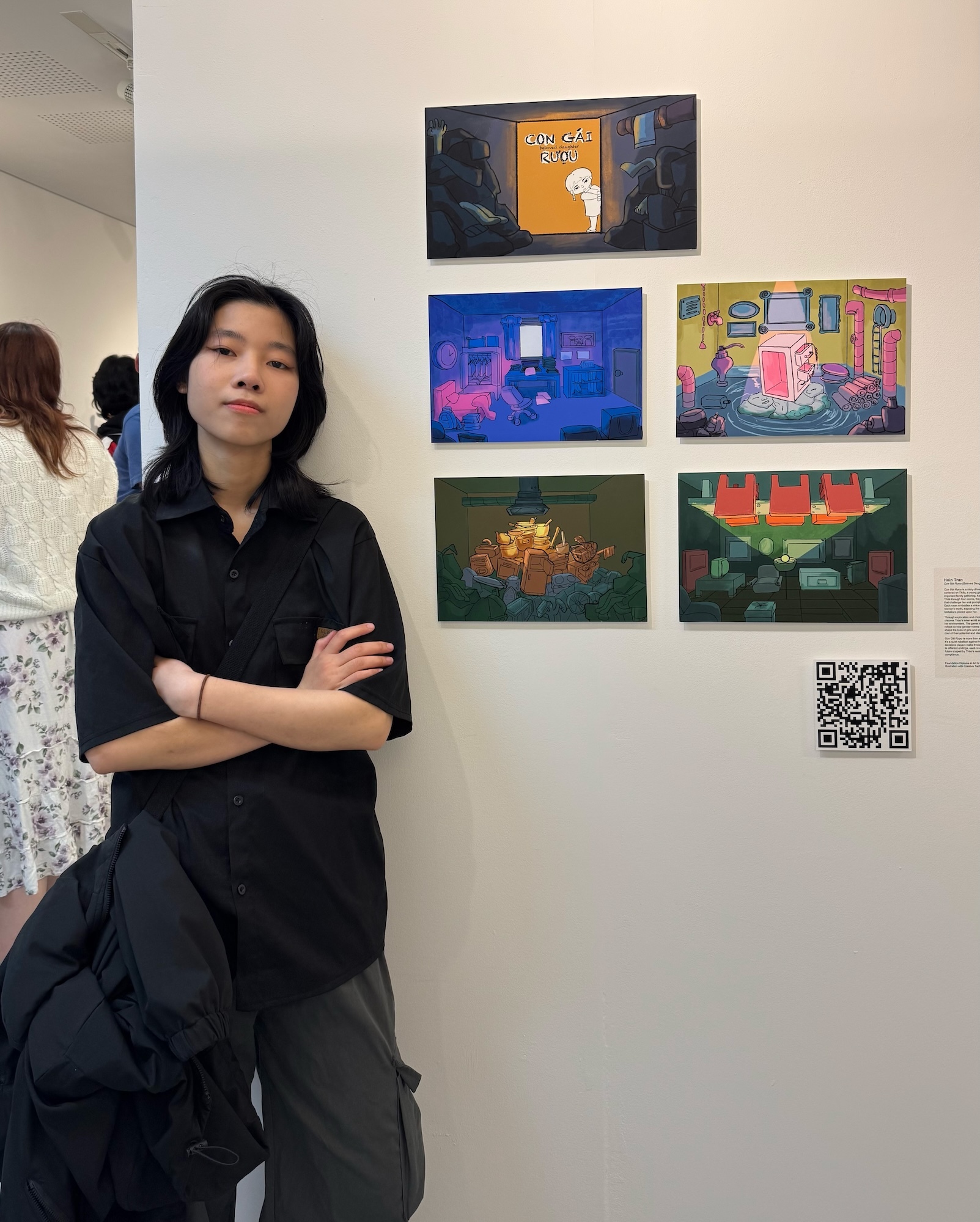 Person in black outfit with crossed arms stands beside wall displaying five colourful digital artworks and QR code in gallery setting.