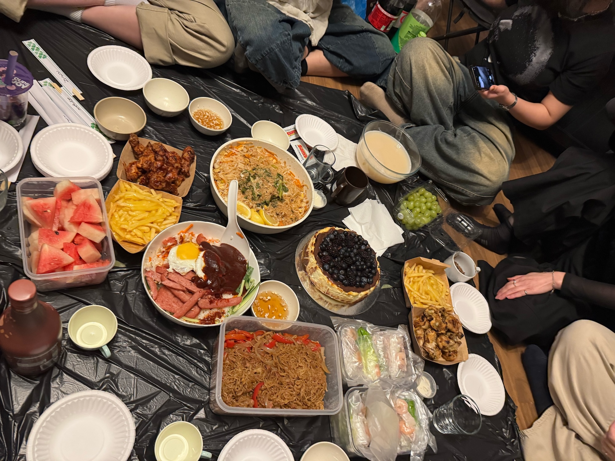 People seated around table laden with various dishes including watermelon, rice, beans, meat, and other foods in bowls and plates.