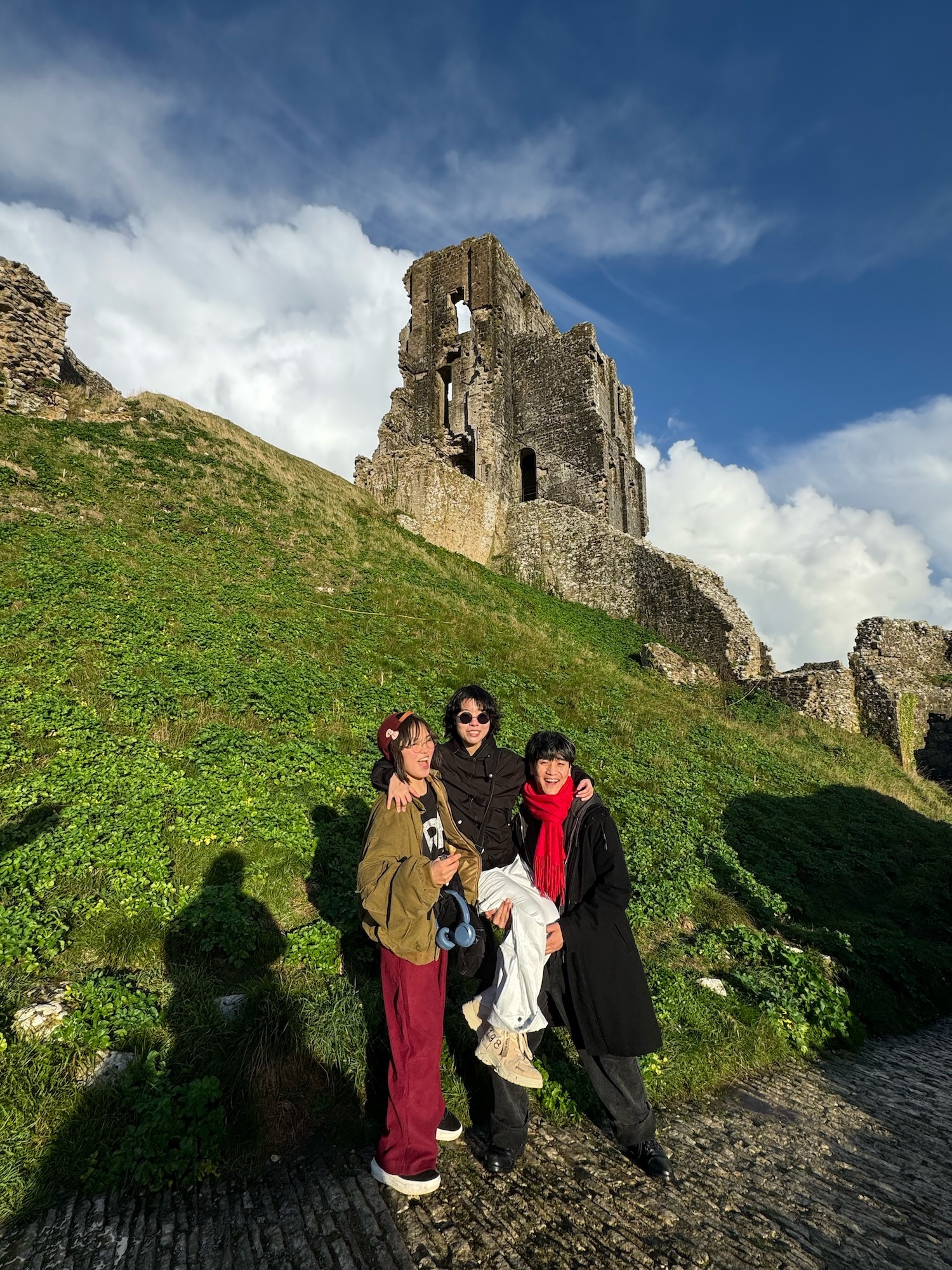 Three people posing on green grassy hill below stone castle ruins against blue sky with white clouds.