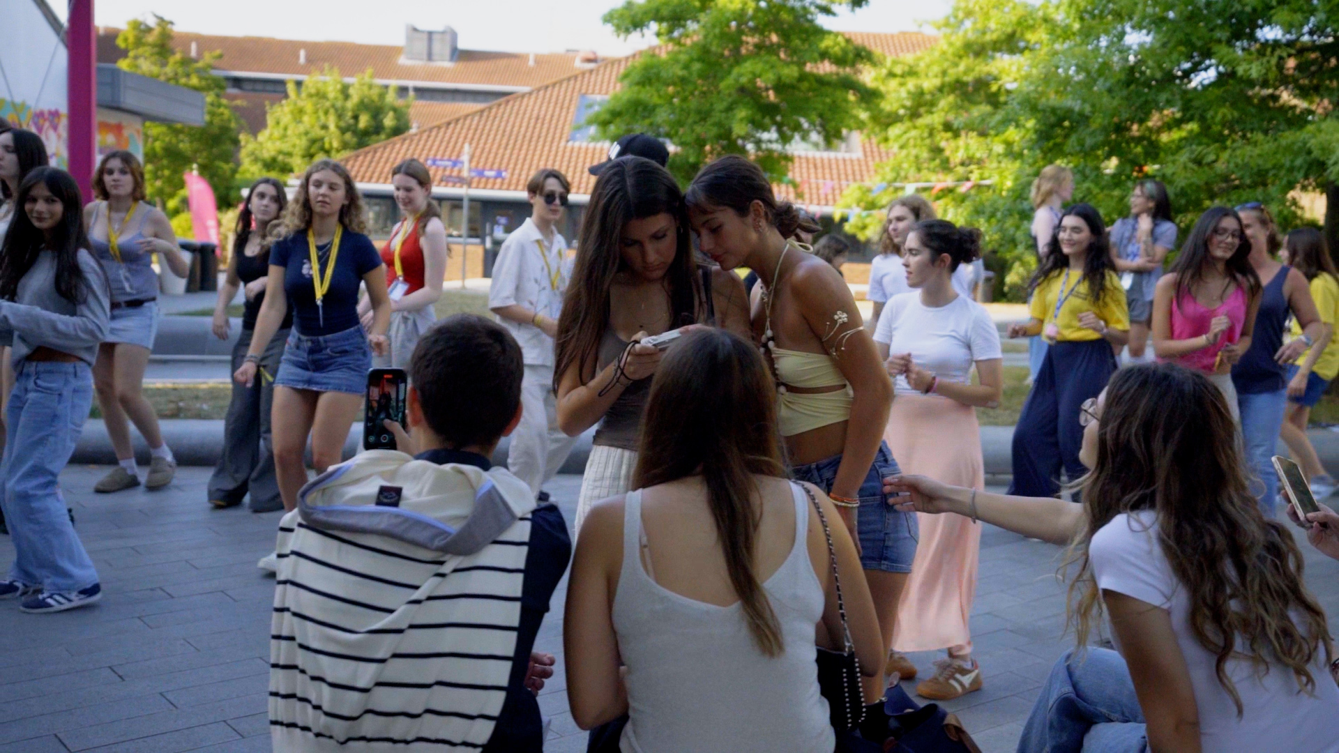 Group of young people gathered in outdoor courtyard with orange-roofed buildings and green trees in background.