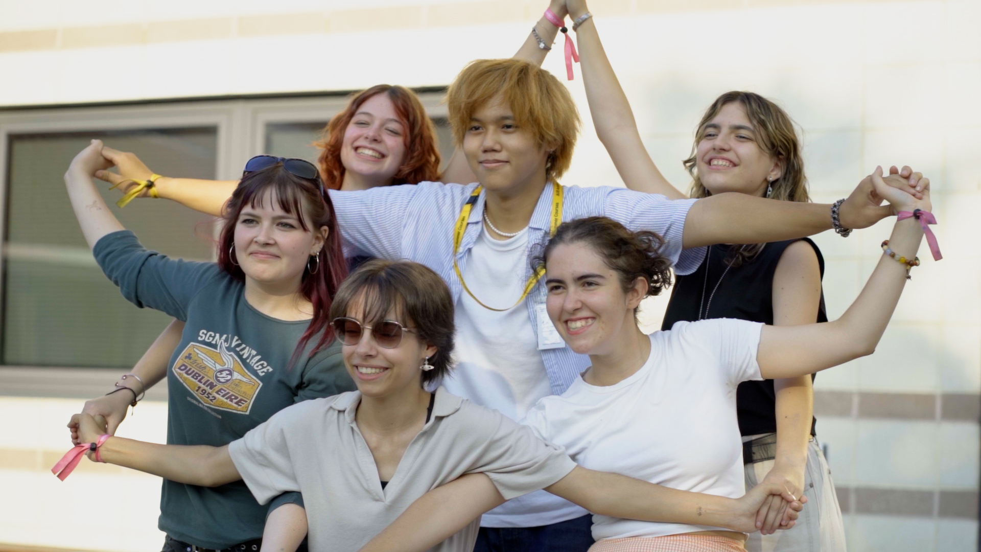 Six young women with raised arms celebrating together outdoors, wearing casual clothing in various colours including white and green.