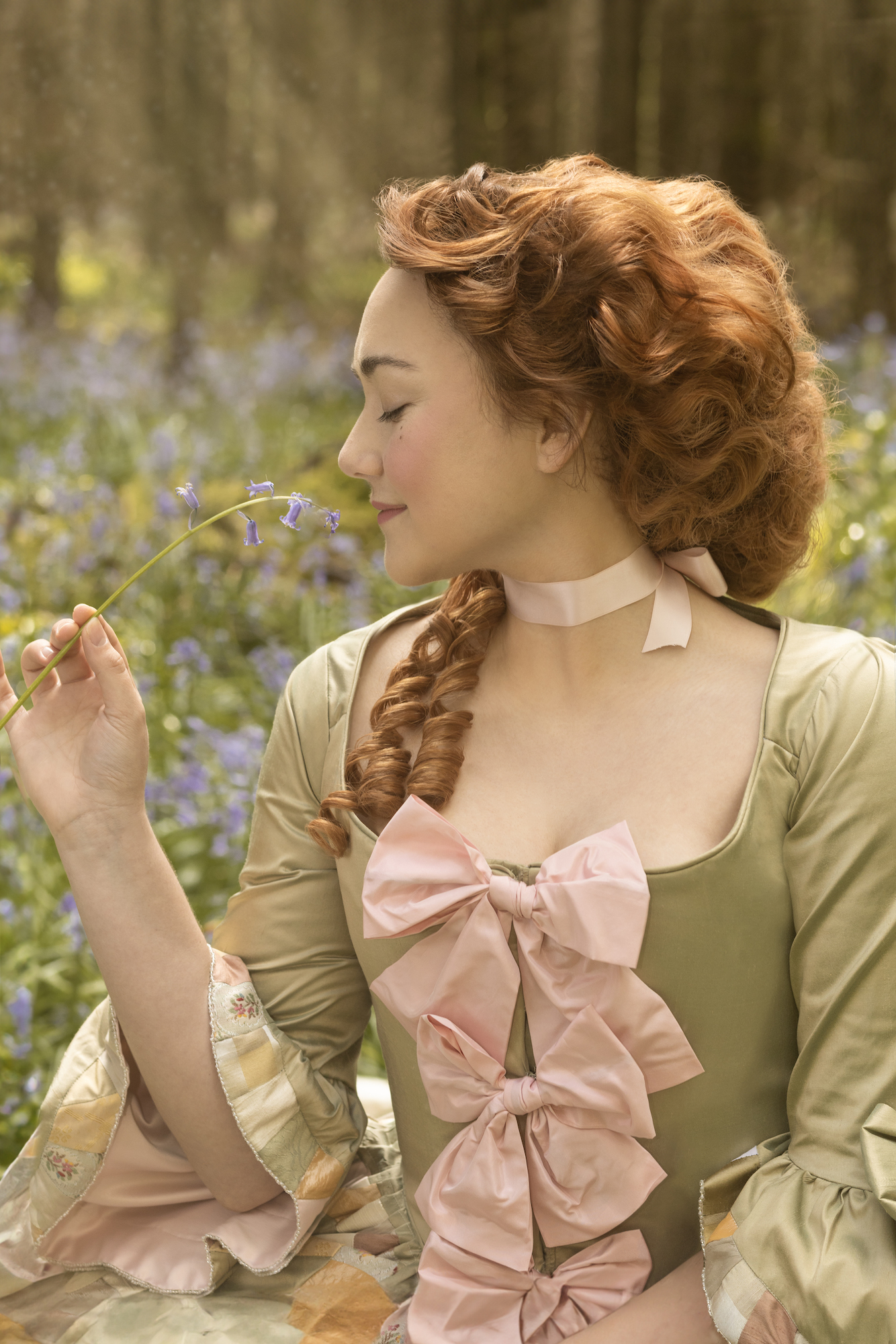 A woman in a period costume sits in a woodland, holding a stem of bluebells and smelling it.