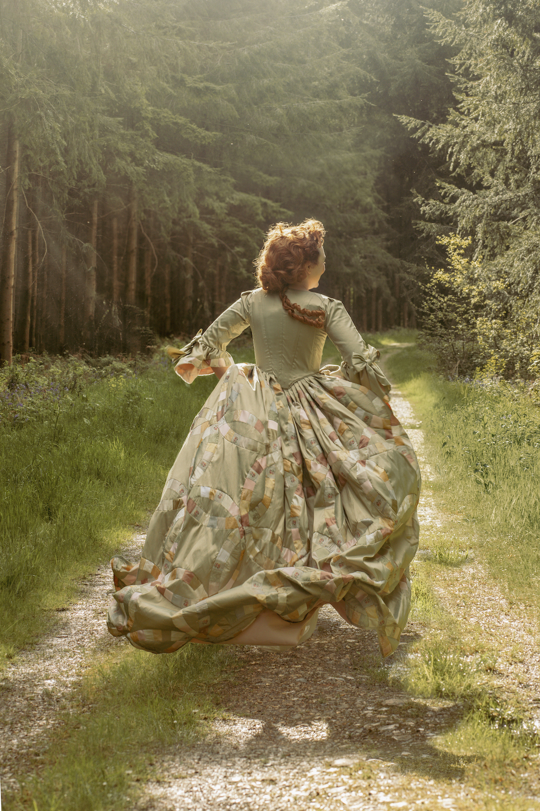 A woman in a period gown running down a grass and gravel path towards a woodland.
