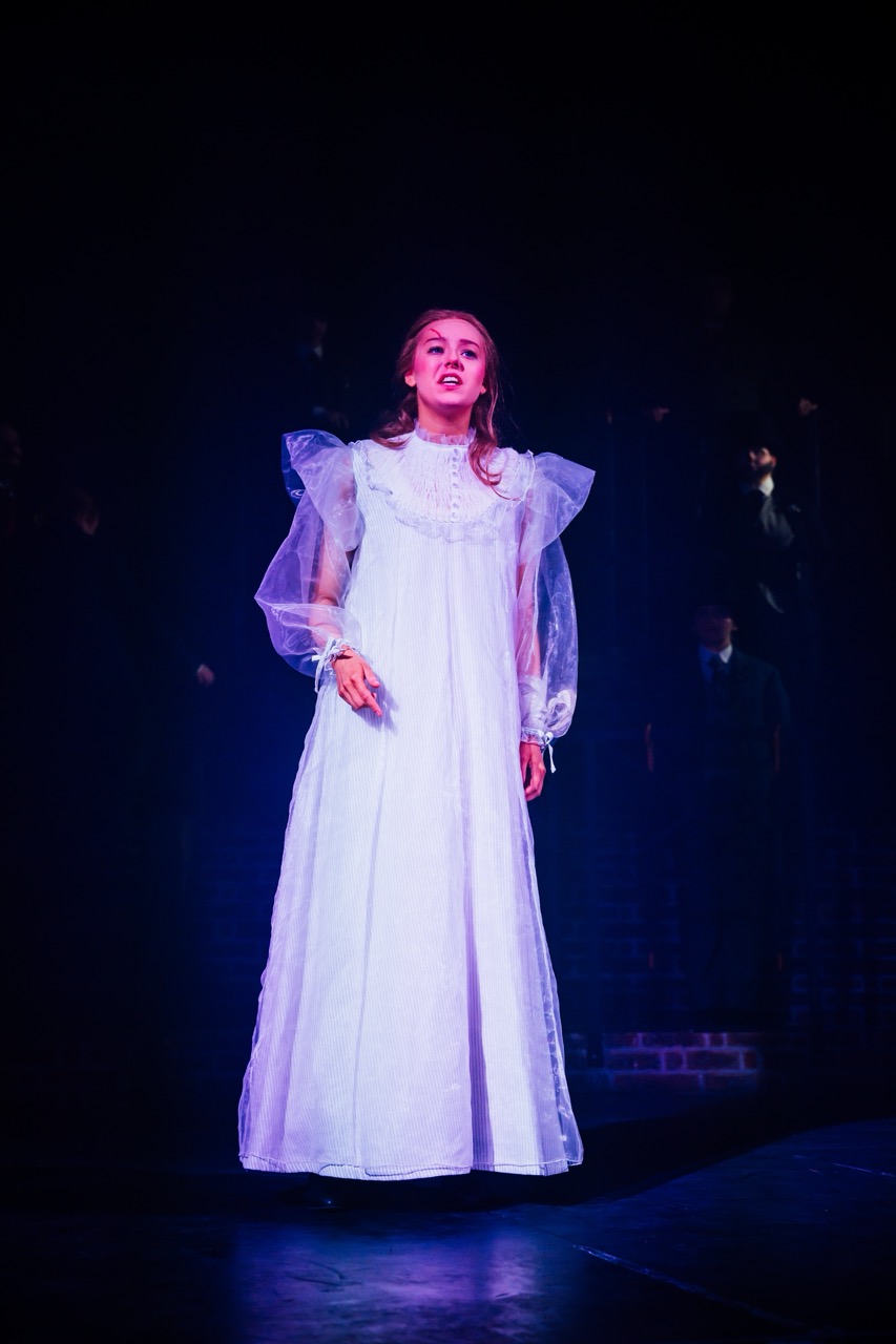 A woman wearing a long white dress with puffy sleeves stands in a dimly lit space. The background features indistinct figures, suggesting a theatrical performance setting.