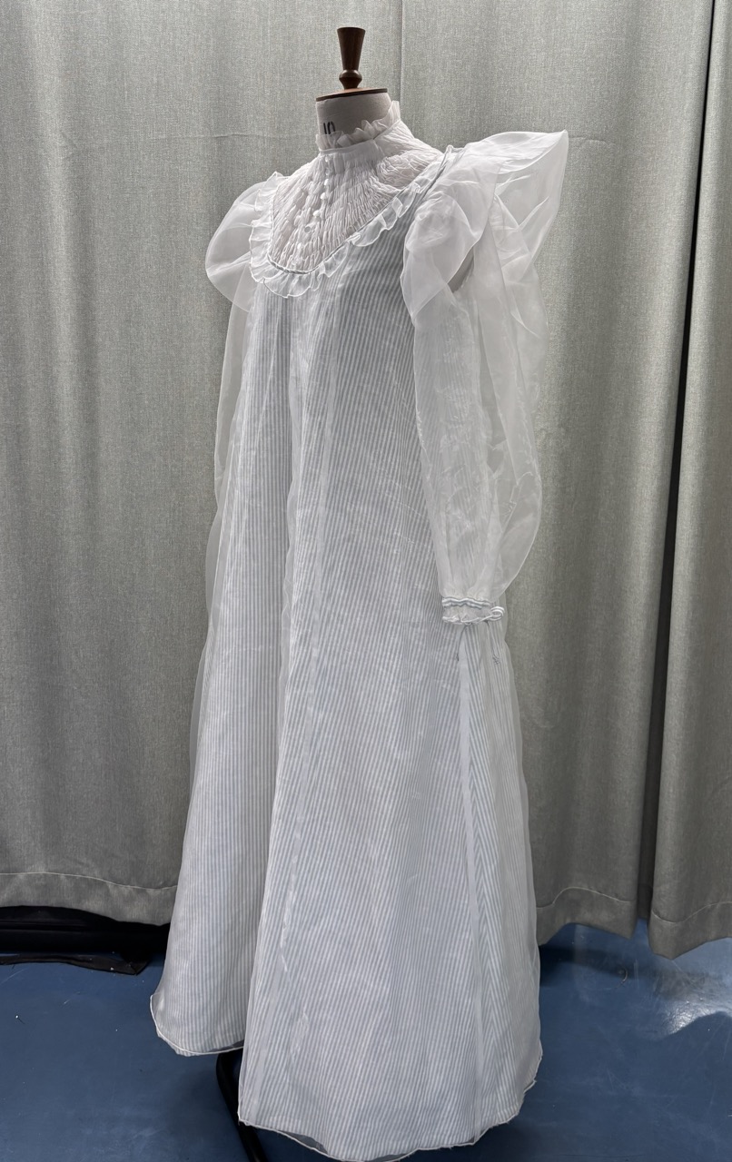 A full-length white dress displayed on a dress form. The dress features puffy sleeves, a high neck with ruffles, and a full skirt. It is set against a backdrop of grey curtains and a blue floor.