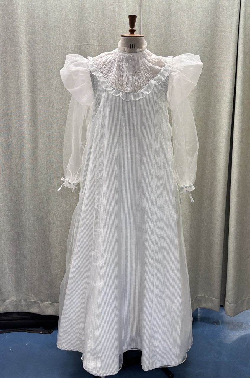 A white dress displayed on a mannequin, featuring a high ruffled collar and puff sleeves. The garment has a floor-length design and is made from a lightweight, sheer fabric, with vertical stripes visible throughout.
