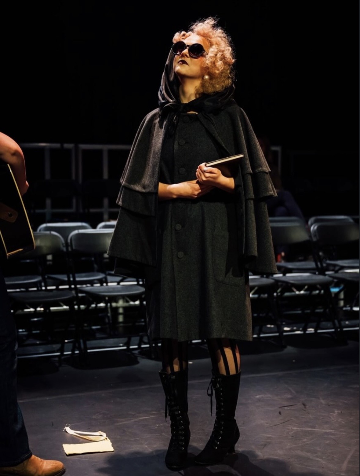 A woman with curly blonde hair, wearing sunglasses and a black cape, stands with her head tilted upwards. She is holding a book in her hand, and black lace-up boots are visible. The background features dark stage lighting with empty chairs.