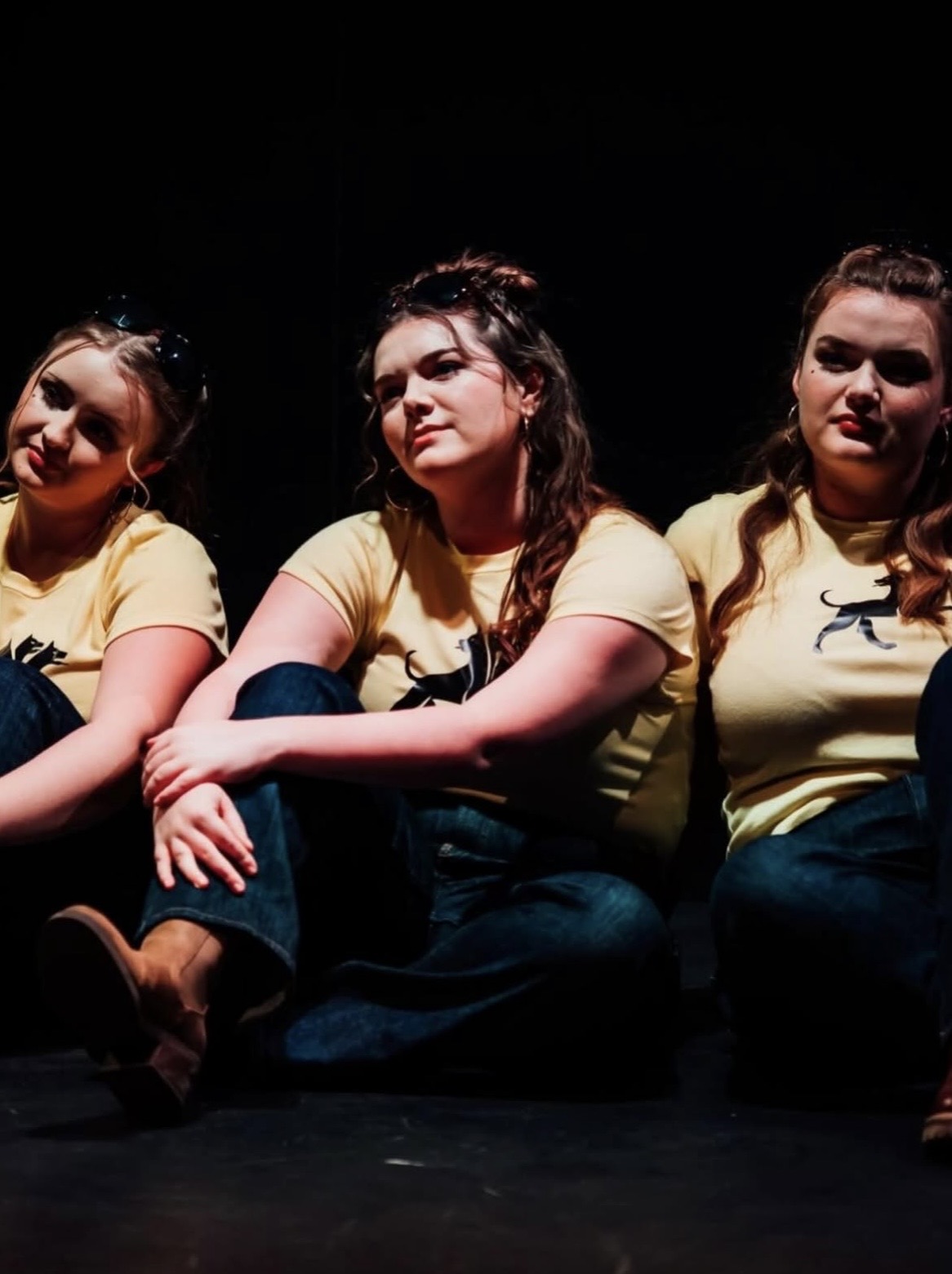 Three women sit side by side on a dark stage. Each woman wears a yellow t-shirt featuring a black cat design, paired with blue jeans. The background is dimly lit, highlighting their coordinated outfits.