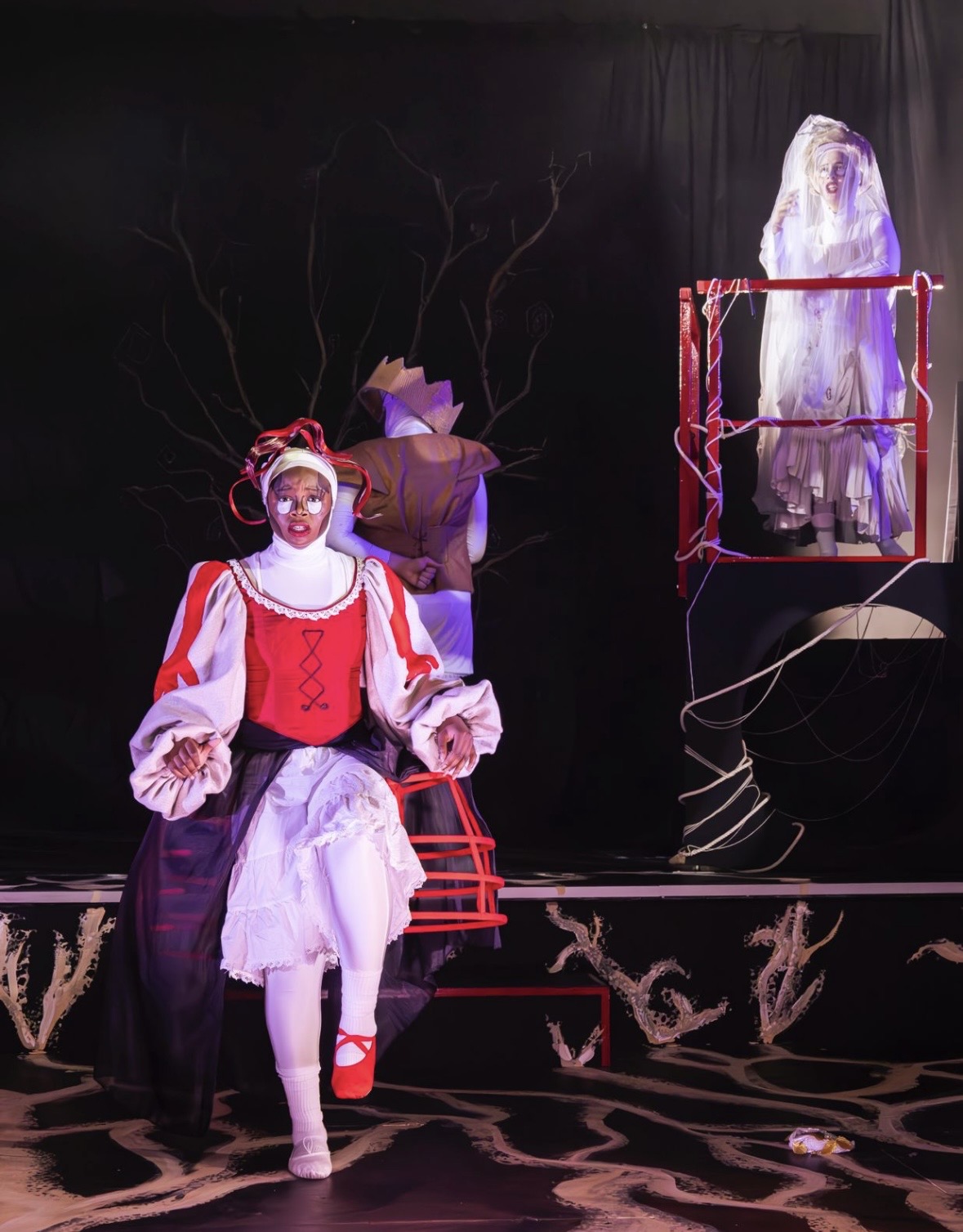 A theatrical scene featuring a woman in a red and white costume with a wide skirt, standing in the foreground. Behind her, a ghostly figure in white is suspended in a red frame, while another character in brown is partially visible in the background. The stage is dimly lit.