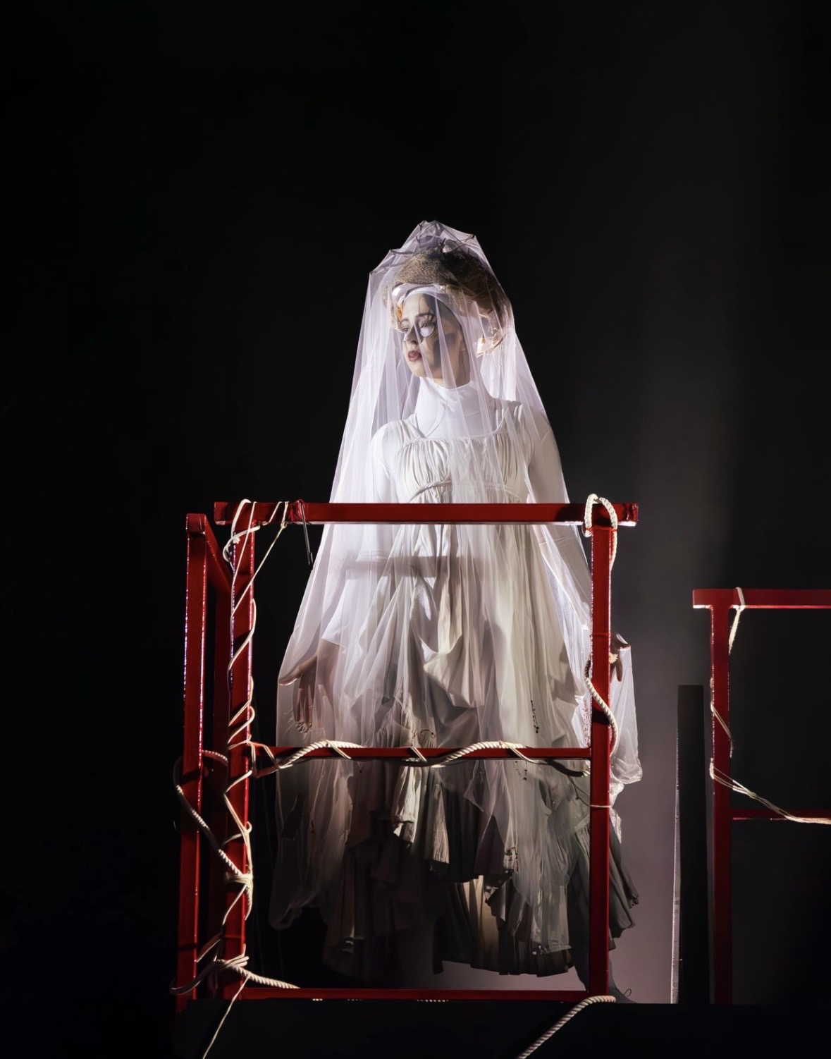 A woman dressed in a flowing white gown and veil stands on a red structure, partially shrouded in fog. The scene is dimly lit, highlighting the delicate layers of her outfit against a dark background.