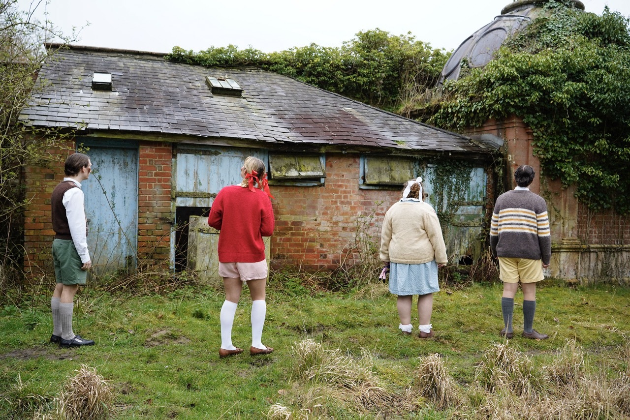 Four characters dressed as children from the 1940s standing in front of a derelict building.