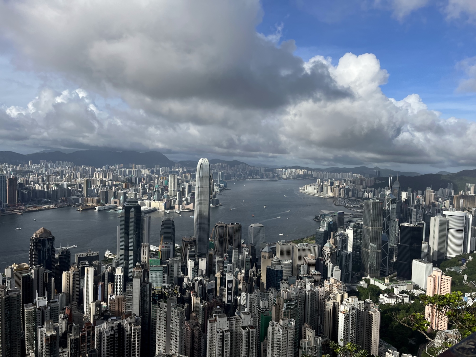 A panoramic view of a dense city skyline beside a wide harbour, with high-rise buildings, distant hills and large clouds overhead.