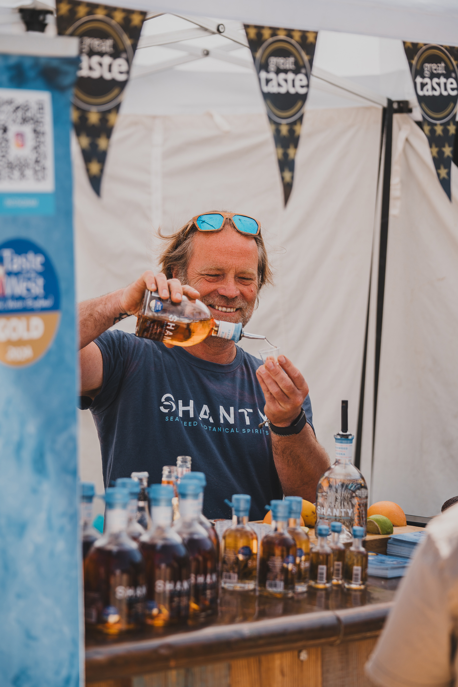 Hugh Lambert smiling while pouring rum into a shot glass, multiple bottles in front of him under a white marquee.