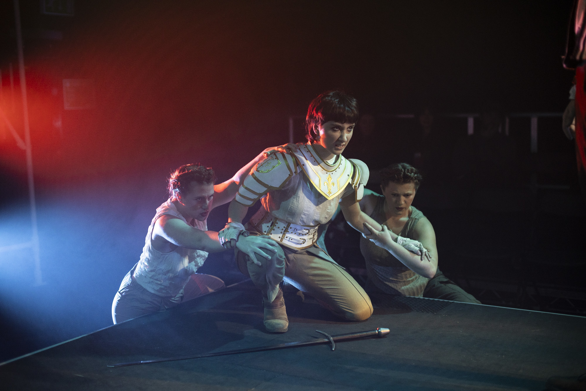 Three BA (Hons) Acting students performing "I, Joan". One of the performers is kneeling on the stage wearing armour, held back by the other two actors standing behind them.