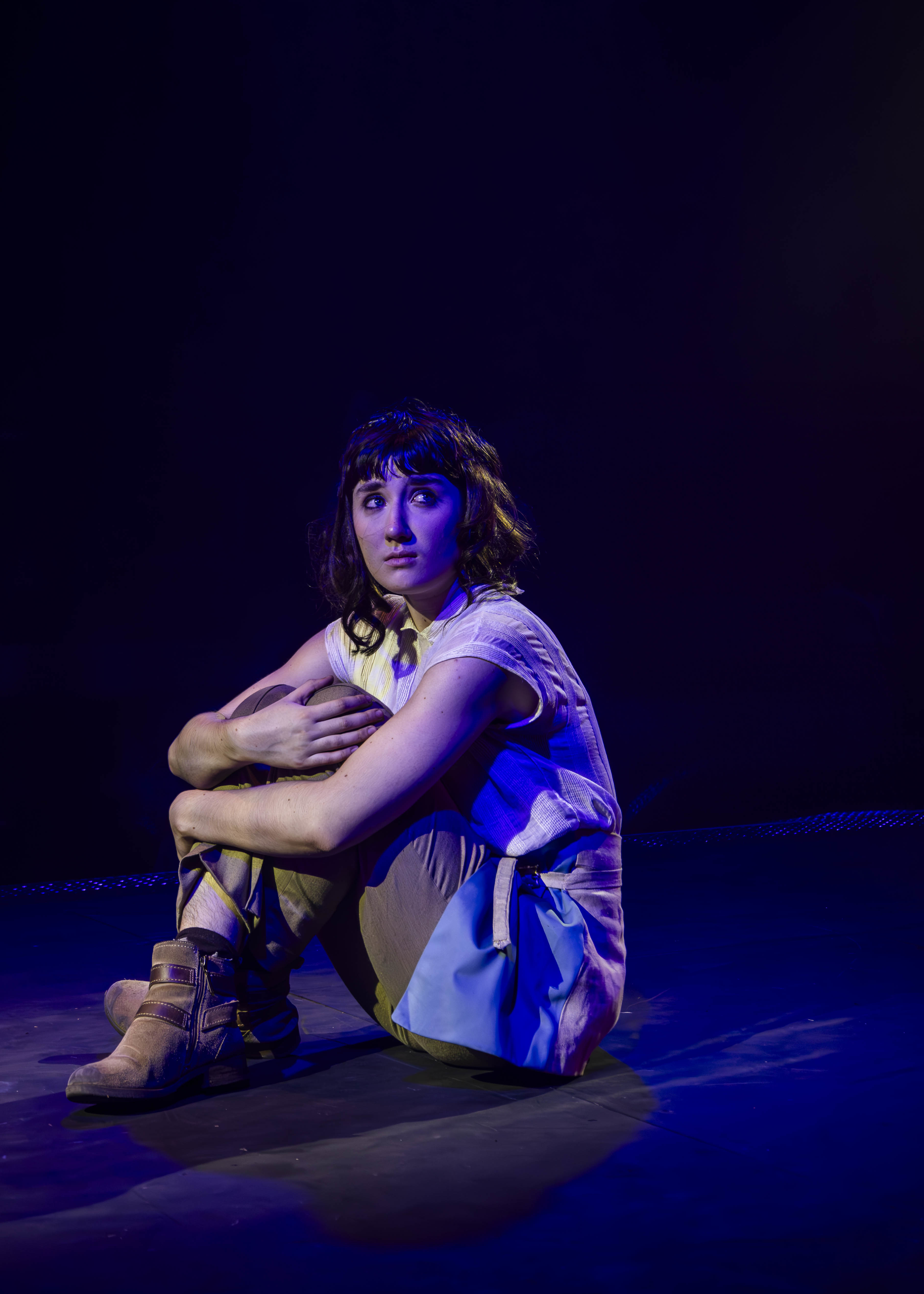 A BA (Hons) Acting student performing I, Joan. They are sitting on stage under a dim blue light, looking emotionally up away from the camera, their arms around their knees.