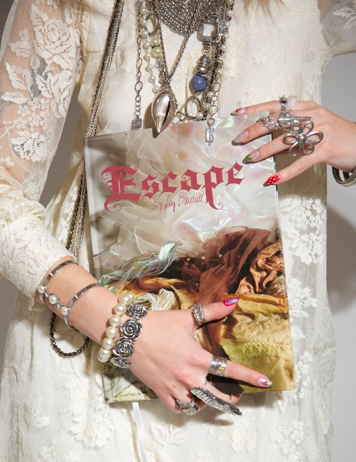 Hands wearing multiple silver rings and bracelets over white lace fabric, with layered necklaces and red gothic text overlay.