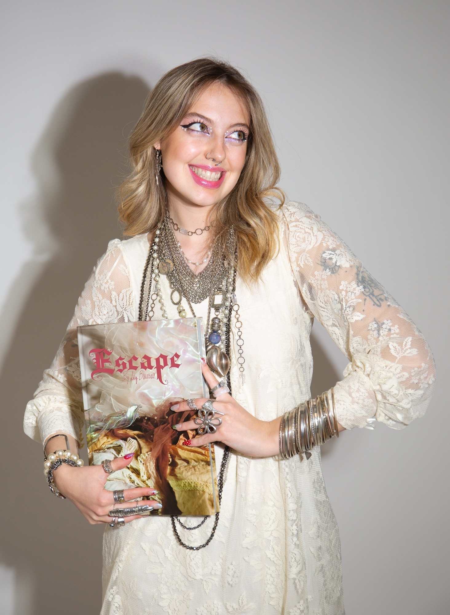 BA (Hons) Fashion Communication student L B Wyatt in white lace dress with layered necklaces and bracelets, holding a copy of her Final Major Project, "Escape", smiling and looking to the side against grey wall.