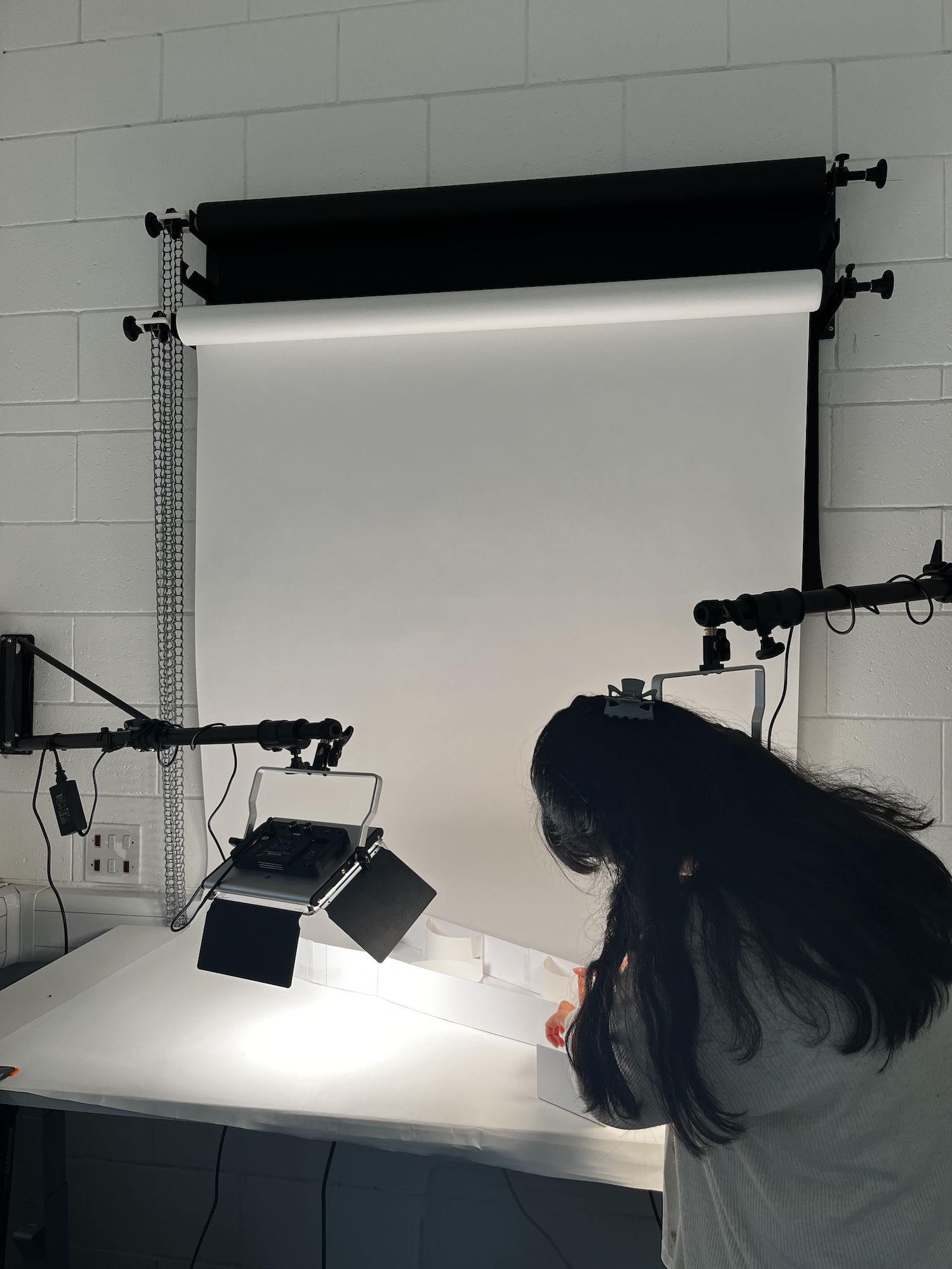 BA (Hons) Interior Architecture and Design student working at photography studio setup with white backdrop, lighting equipment, and copy stand against white brick wall.