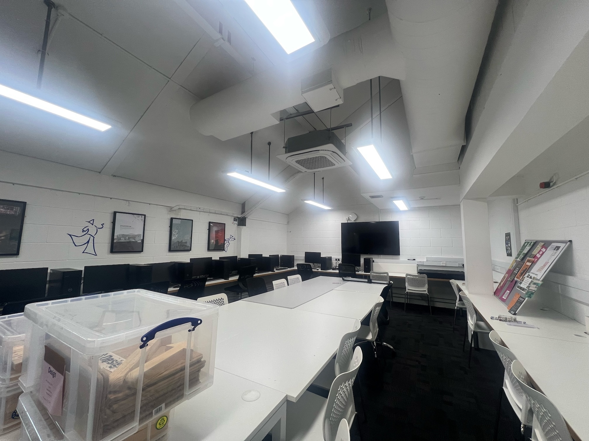 Empty computer suite with white walls, fluorescent lighting, rows of black chairs, wall-mounted TV, and storage boxes in foreground.