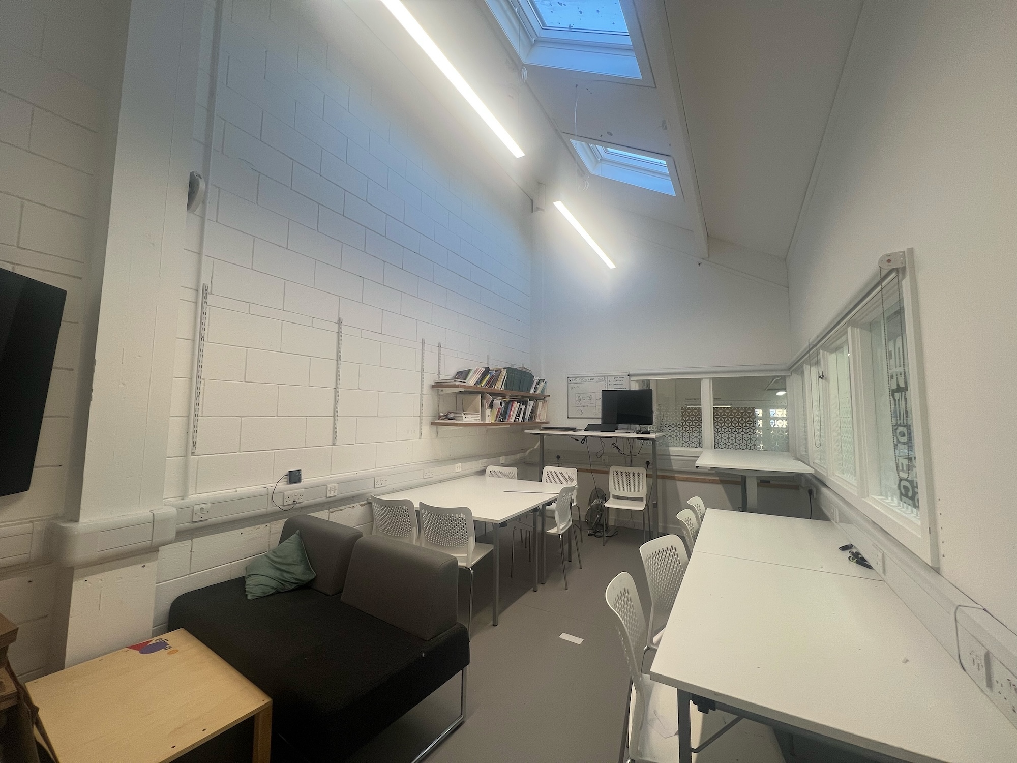 Modern white office space at Arts University Bournemouth with sloped ceiling, skylights, LED strip lighting, grey sofa, white dining table with chairs, and glass railing.