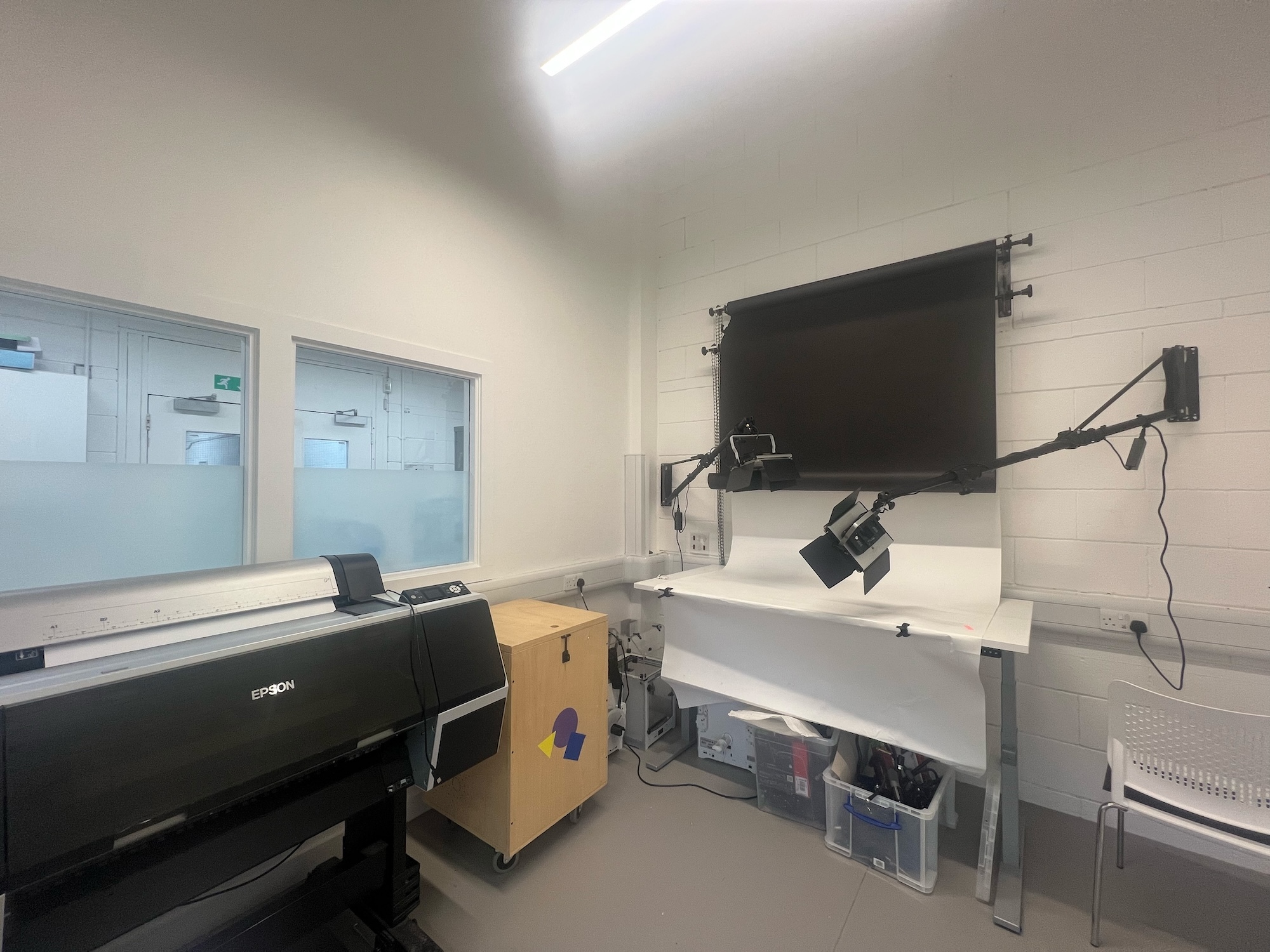 Modern office space with white walls, large windows, white desk, black printer, wooden cabinet, and wall-mounted television.