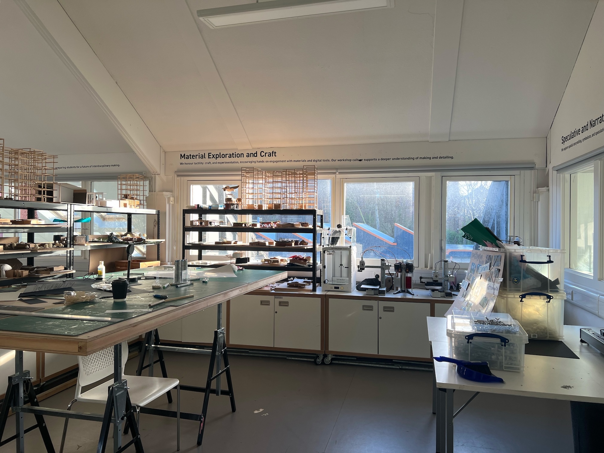 Laboratory with white walls, metal work benches, shelving units with materials, large windows, and equipment under bright fluorescent lighting.