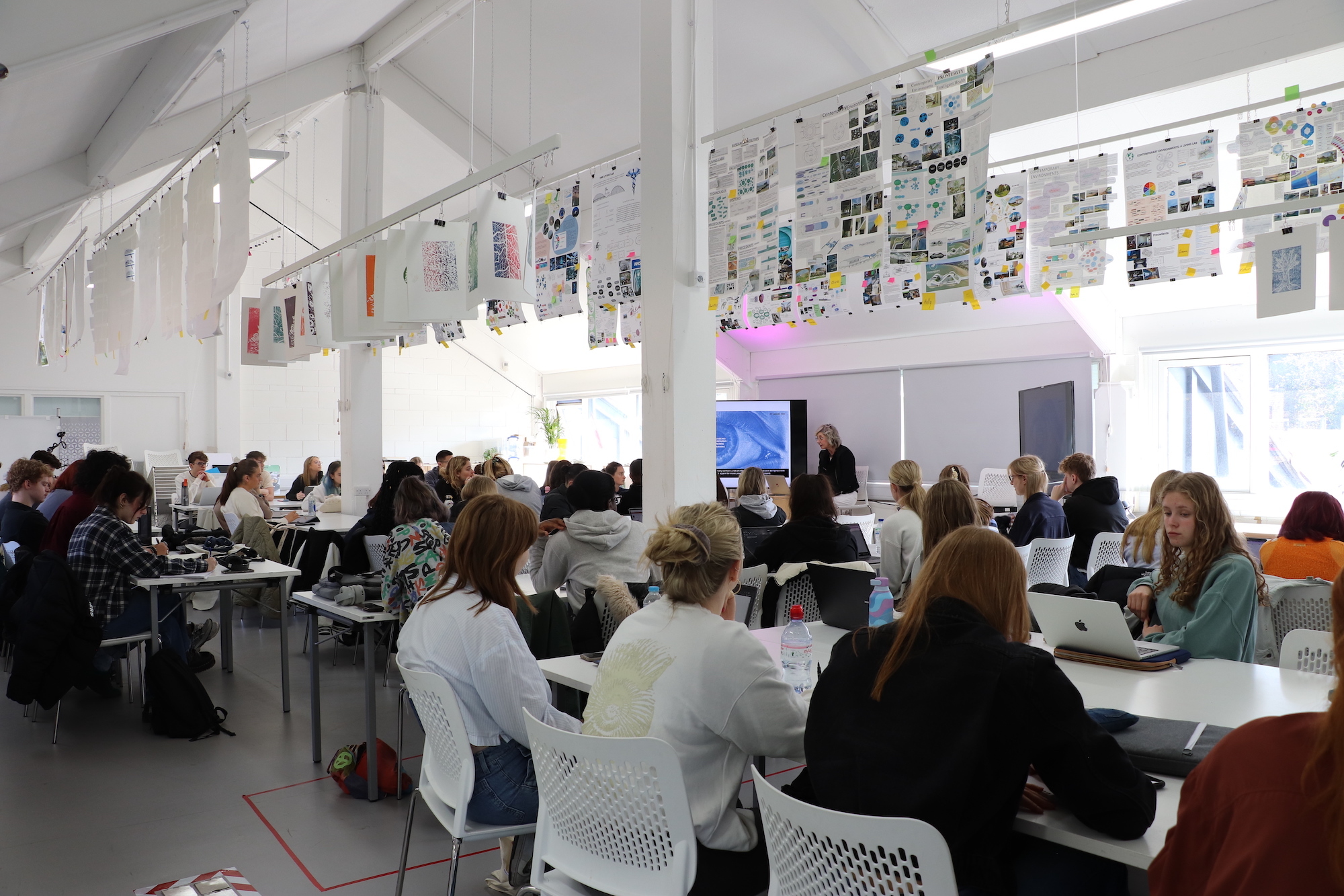 A large group of people sitting at tables in a studio space while a person delivers a lecture.