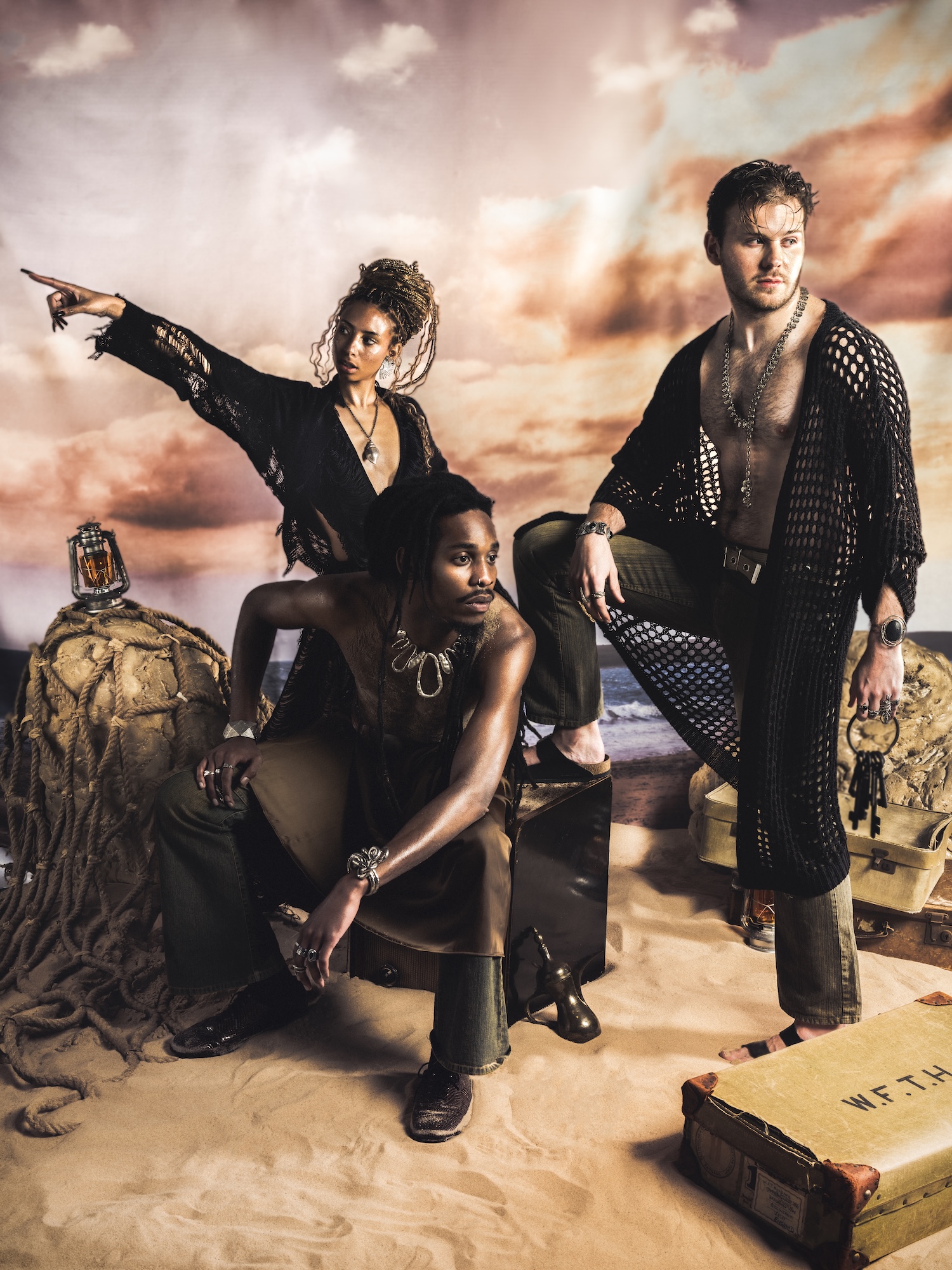 Three models in stylish outfits pose in a desert-themed studio setting. The woman on the left wears a black sheer top and points her right arm, while the man in the centre wears a sleeveless garment. The man on the right is dressed in a mesh top.