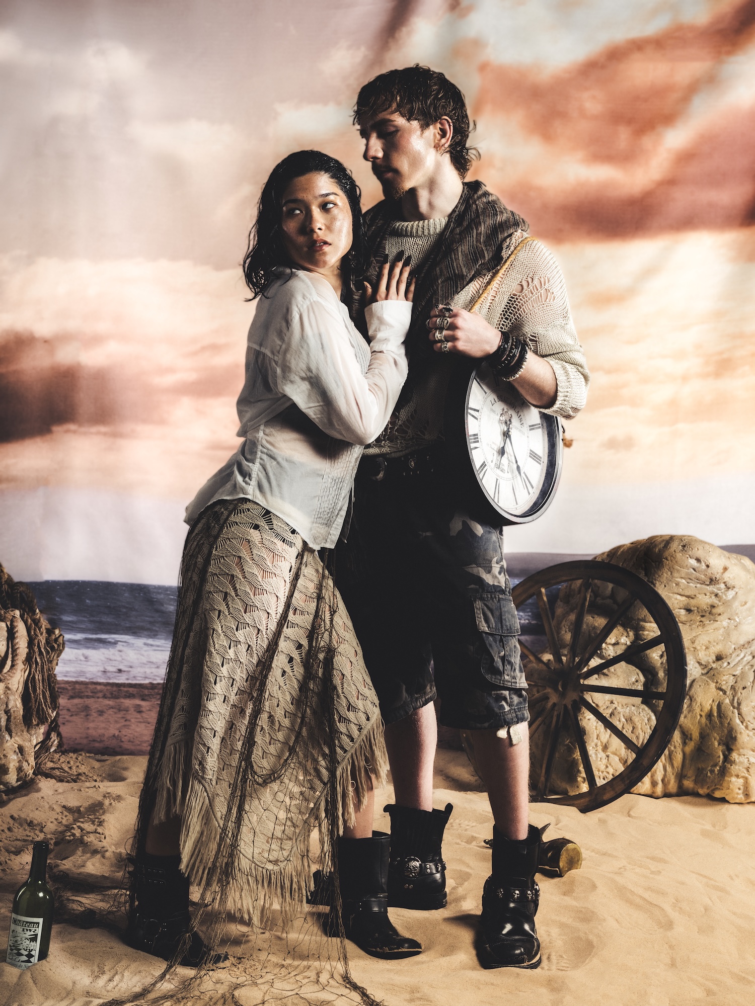 A man and a woman pose against a backdrop depicting a beach scene with a sunset. The woman wears a light blouse and long skirt, while the man is dressed in a knit sweater and cargo shorts, holding a large clock. They stand near a wooden cart on the sand.