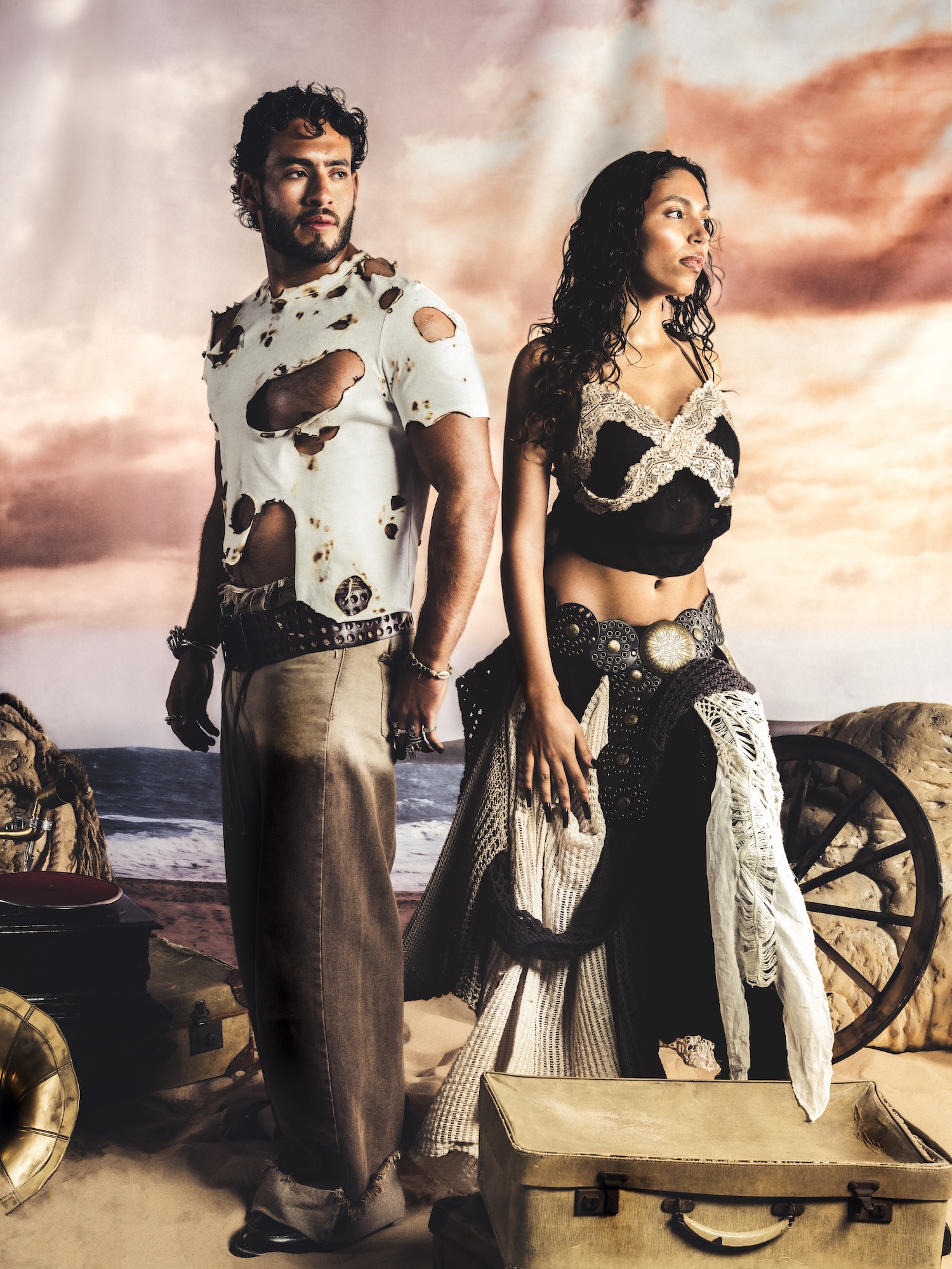 A man wearing a torn white shirt and grey trousers stands next to a woman in a black top and white skirt. They are posed against a backdrop of a beach scene with clouds, surrounded by various props, including a cart and a suitcase.