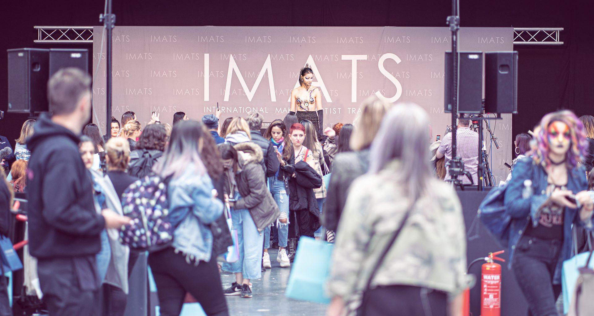 A crowd of people walking around a stage. A performer stands on stage. The word 'IMATS' is behind them on a banner.