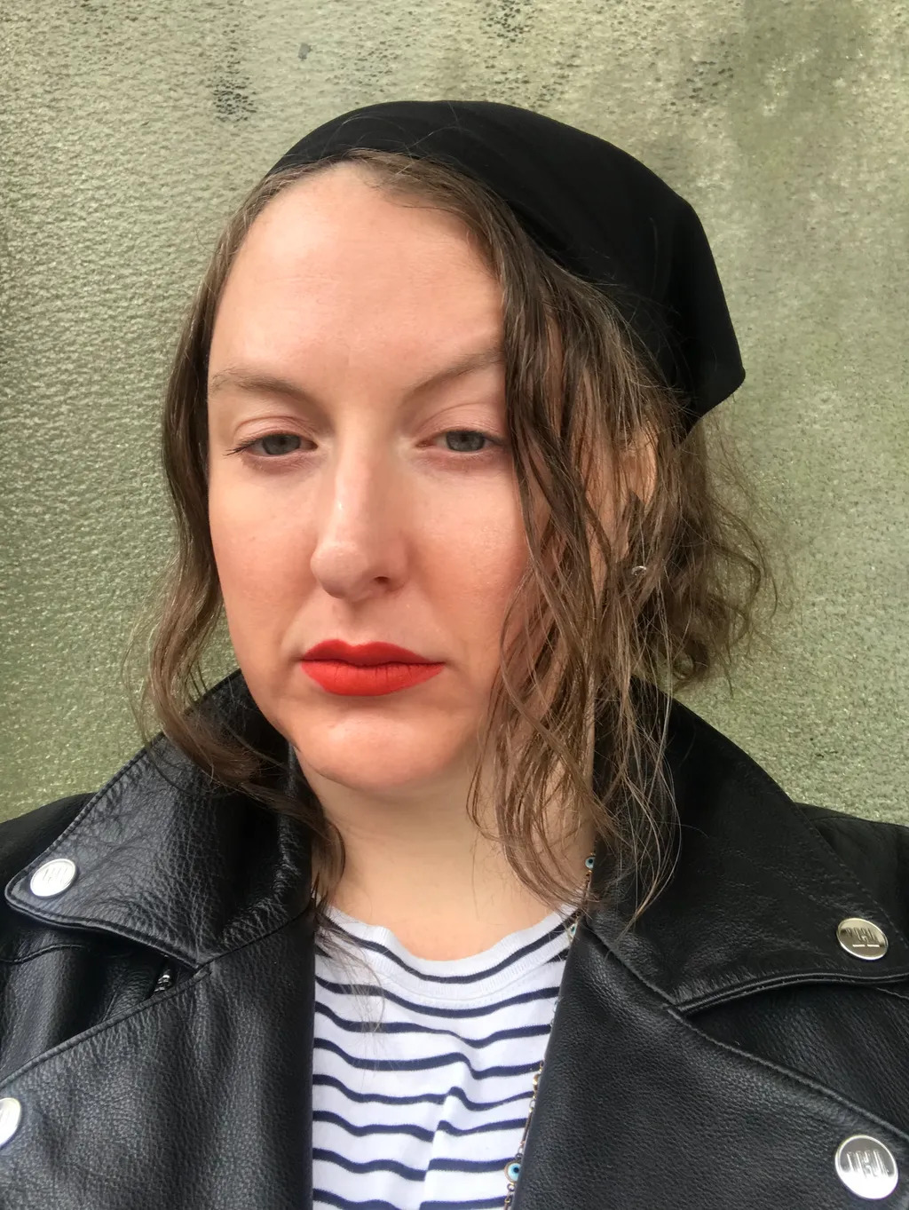 Woman wearing black beret and leather jacket with striped top, red lipstick, wavy brown hair, against textured beige wall.