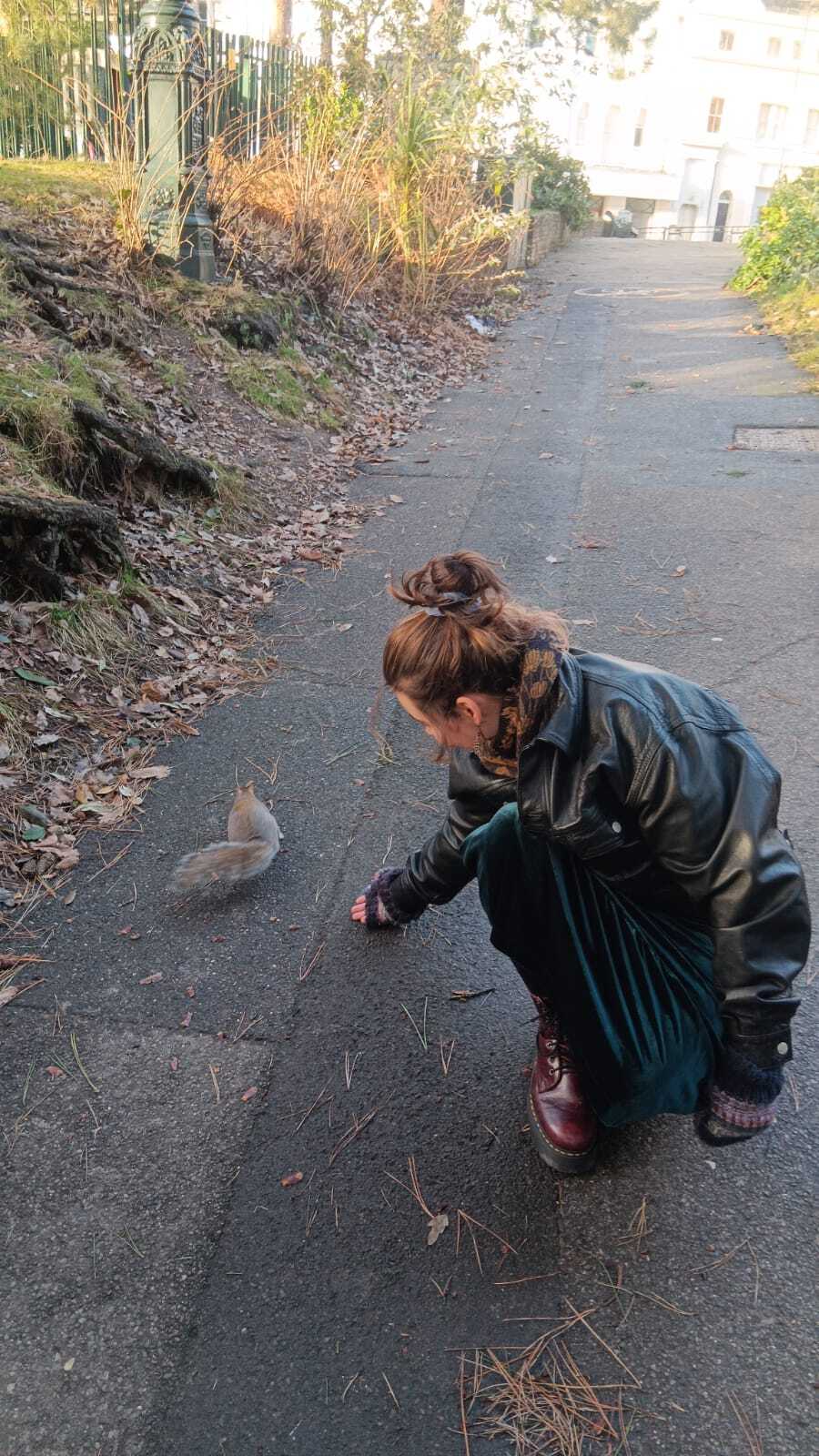 Woman in green jacket crouching on pavement path, extending hand towards a squirrel. Overgrown vegetation and buildings visible.