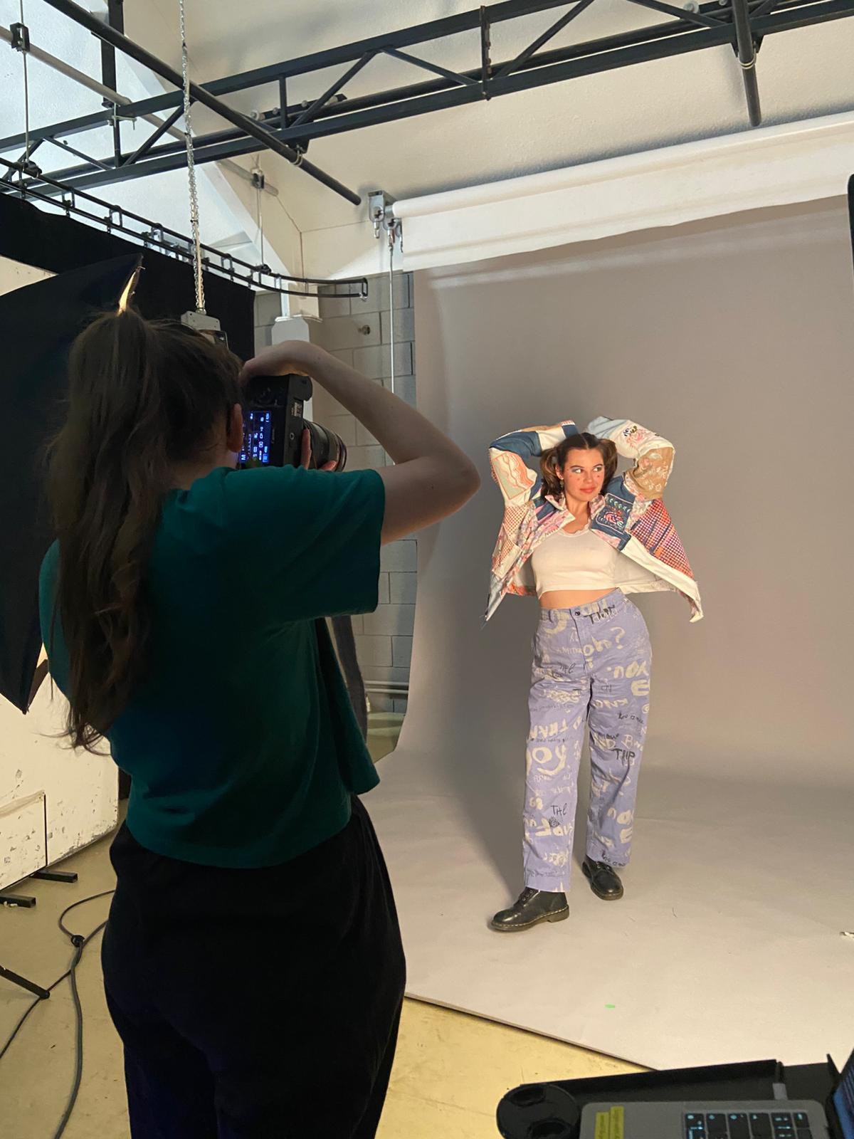 Camera operator filming model posing with arms raised in white studio with exposed ceiling beams and lighting equipment.