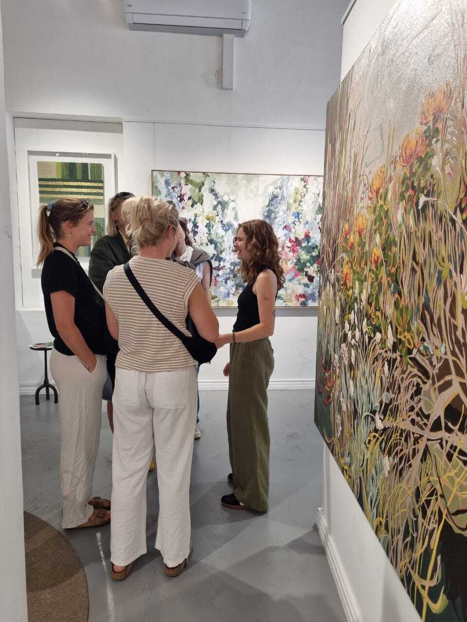 Four people standing in white-walled gallery space viewing colourful paintings, including large floral canvas on right wall.
