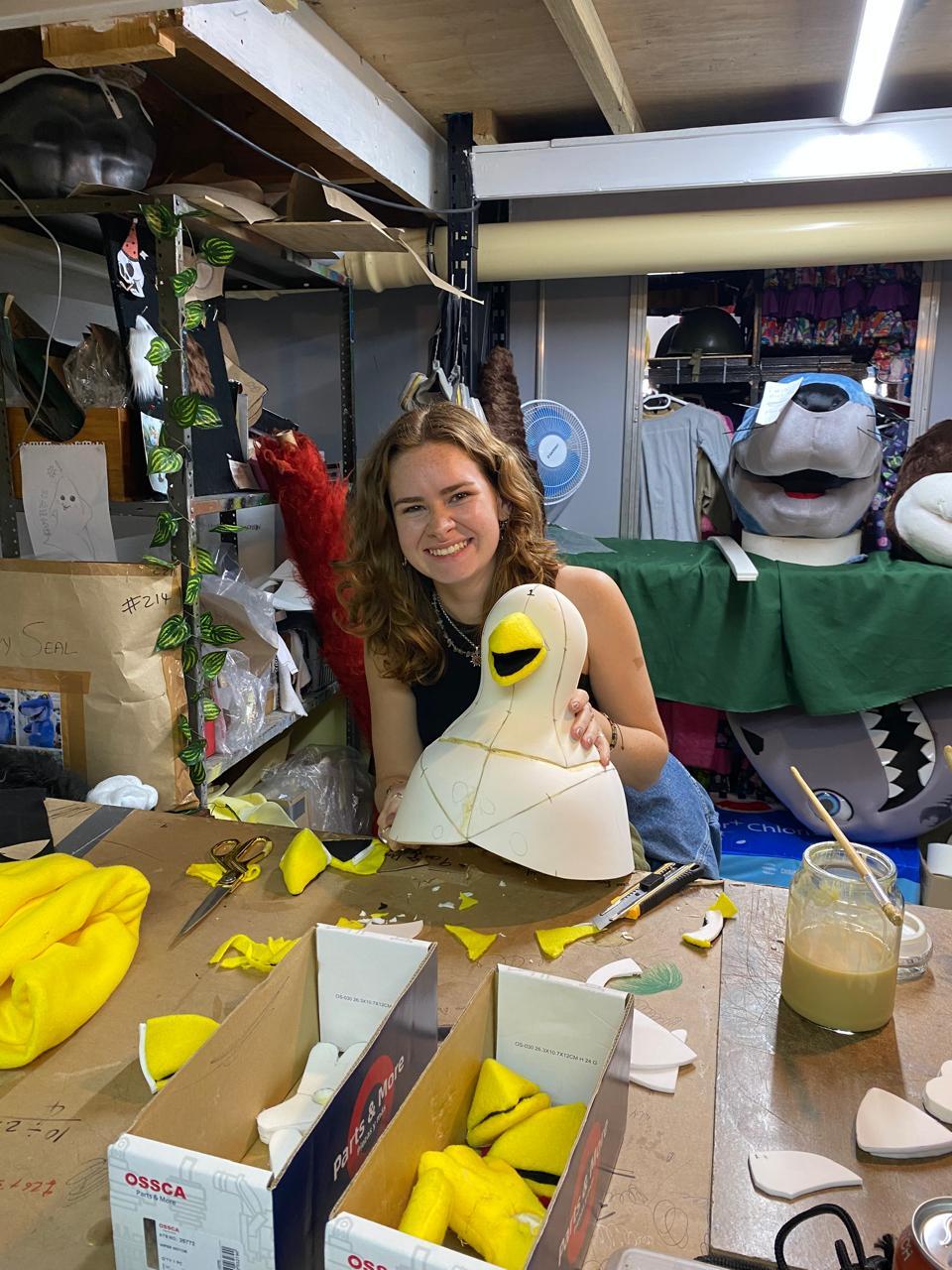 BA (Hons) Textiles Design graduate H Lee in workshop holding white mascot puppet with yellow beak, surrounded by craft materials and puppet heads on shelves