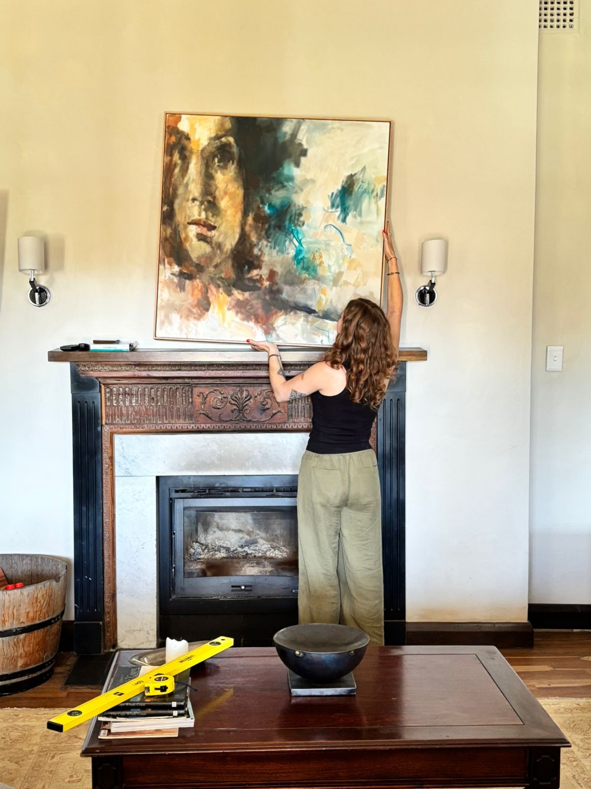 Woman in black top and beige trousers adjusting large abstract painting above ornate wooden fireplace mantel in cream-walled room.