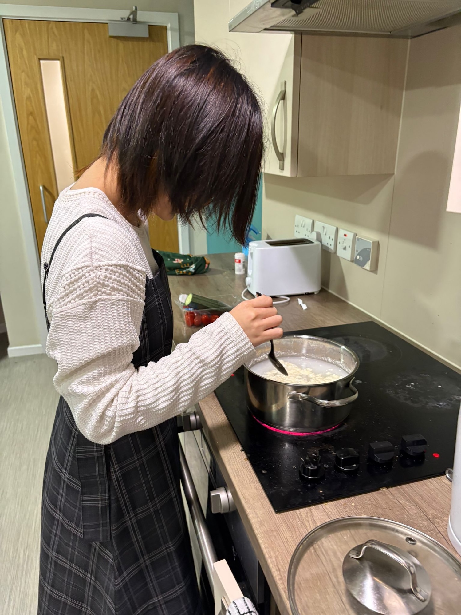 A young person with short hair wearing a white and black checked outfit is stirring a pot on an electric stove in a modern kitchen. The background features cabinets, a kettle, and a work surface with various kitchen items.