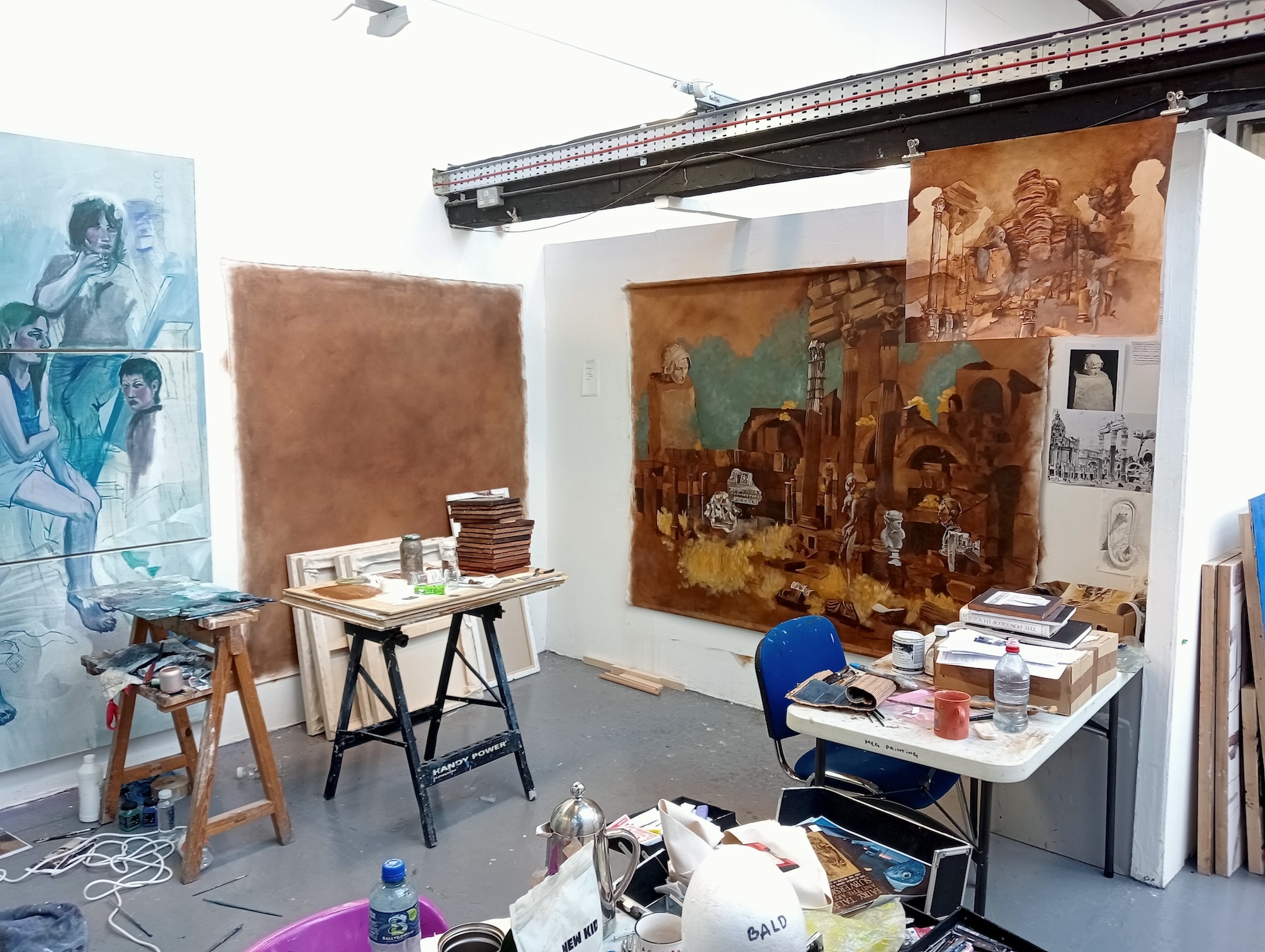 Artist's studio with paintings on white walls, wooden easel, white desk with blue chair, art supplies scattered on floor